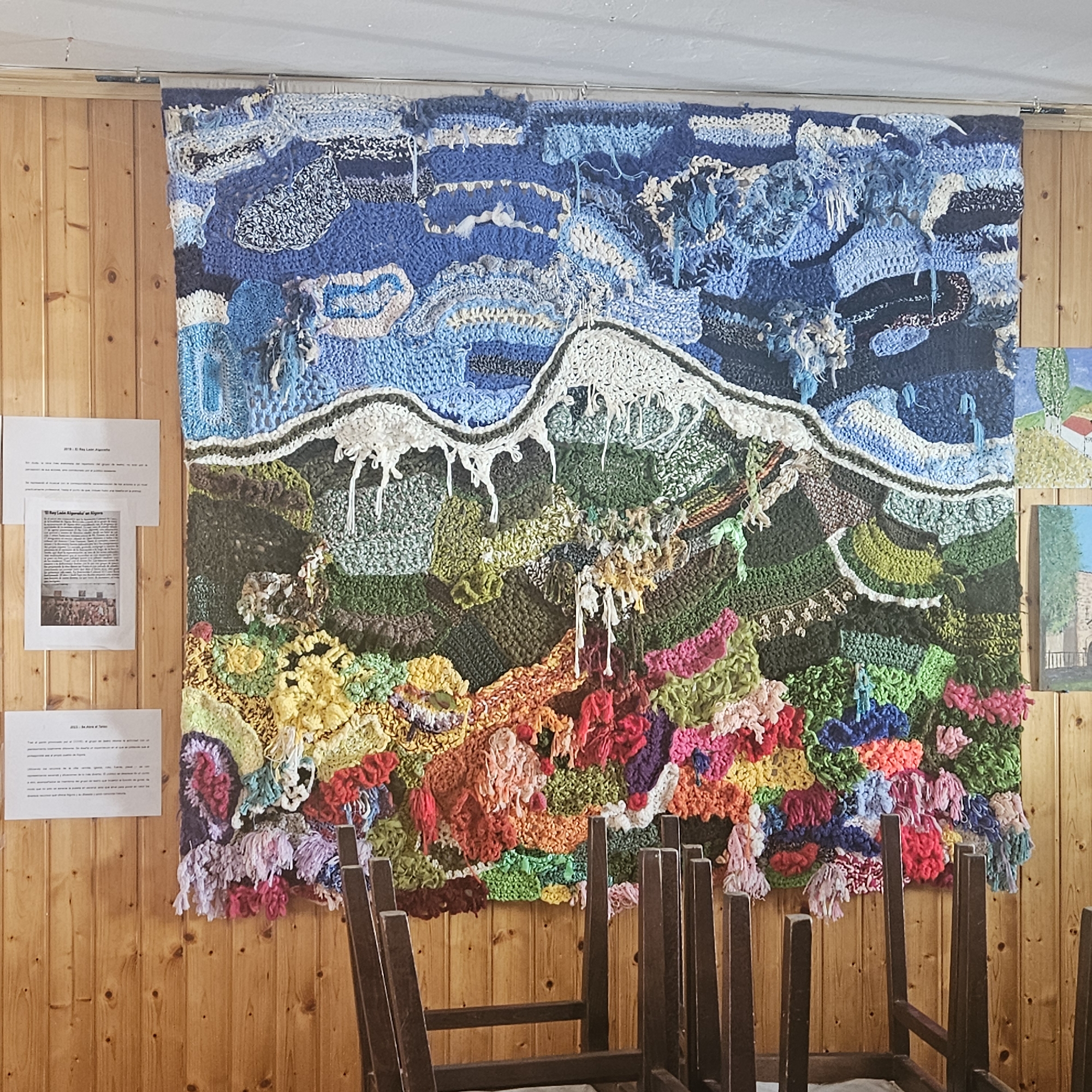 A medium, full shot captures a large, colorful textile wall hanging depicting a mountain landscape in a room with wood-paneled walls. The wall hanging is made of different textures and colors of yarn, creating a three-dimensional effect. The top portion of the hanging shows a blue sky with white clouds, the middle portion a mountain range with snow-capped peaks, and the bottom portion a colorful array of flowers and greenery. To the left of the textile are two sheets of paper with text and images on them. To the right, there are two small paintings. In the foreground, there are several chairs stacked upside down, obscuring the lower portion of the wall hanging. The location is Algora, Spain.