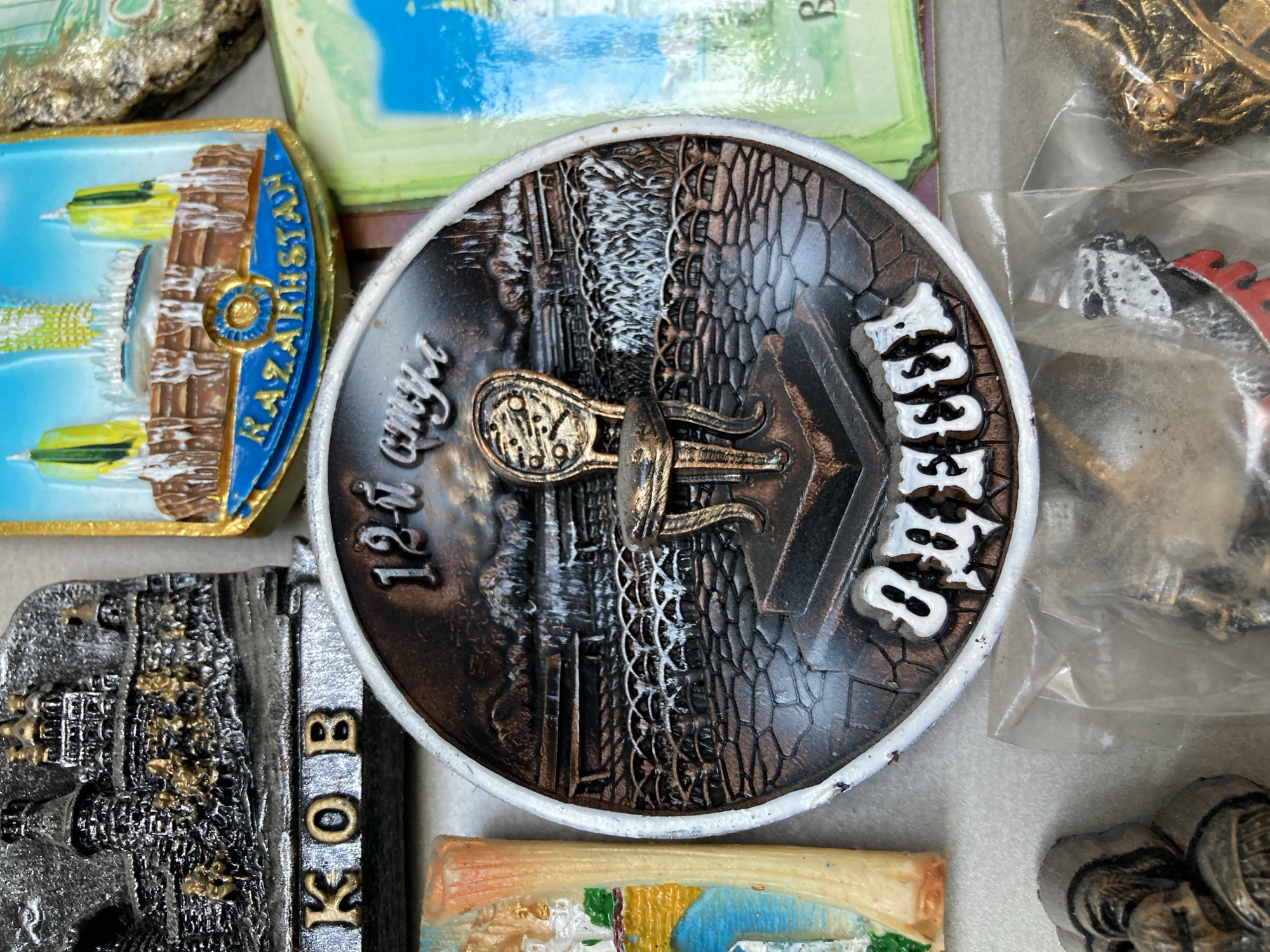 The image displays a collection of souvenir magnets. In the center, a round magnet features a detailed scene with a cobblestone floor, a decorative railing, and a solitary ornate chair positioned on a raised platform. Above the chair, text in a stylized font reads "12-й". The bottom of this magnet has the white text "ОДЕССА". To the left, a rectangular magnet depicts a silver and gold castle with the letters "КОВ" visible below it. To the right of the central magnet, a green magnet shows a stylized landscape with blue water and a white structure. A magnet featuring a building with yellow spires and the word "KAZAKHSTAN" is visible in the upper right portion of the image. Other magnets with abstract designs and metallic figures are also present. The magnets are arranged in a flat lay composition.
