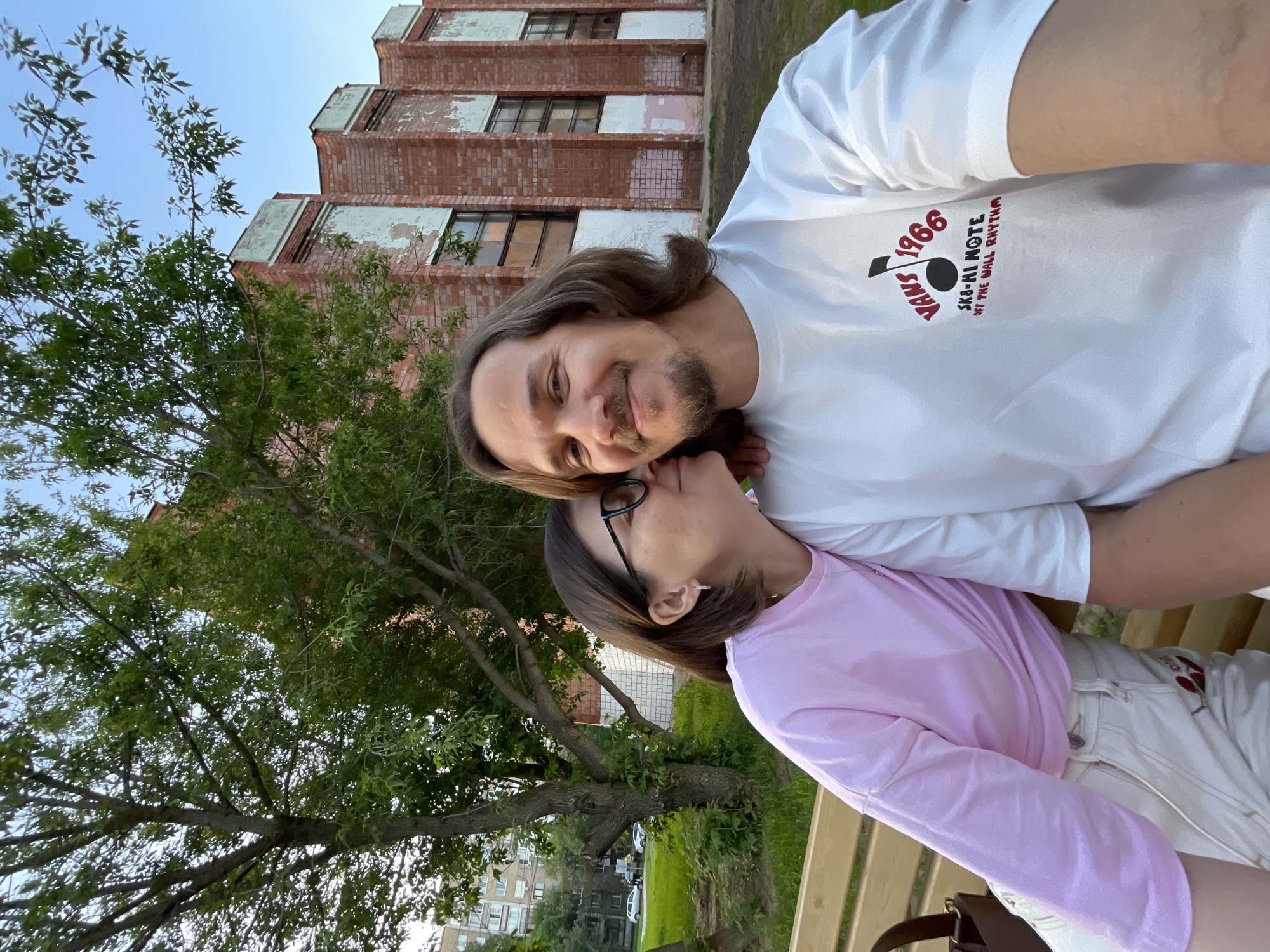 A photo shows a man and a woman in an embrace. They are outdoors, with a tree on the left side of the frame and a brick building in the background.
The man has shoulder-length brown hair and a beard. He's wearing a white t-shirt with a graphic design on it.
The woman has short brown hair and glasses. She is wearing a pink long-sleeved shirt and white jeans.
They appear to be sitting on a bench. The background shows a multi-story brick building with visible weathering and windows. The location is Izhevsk, Russia.