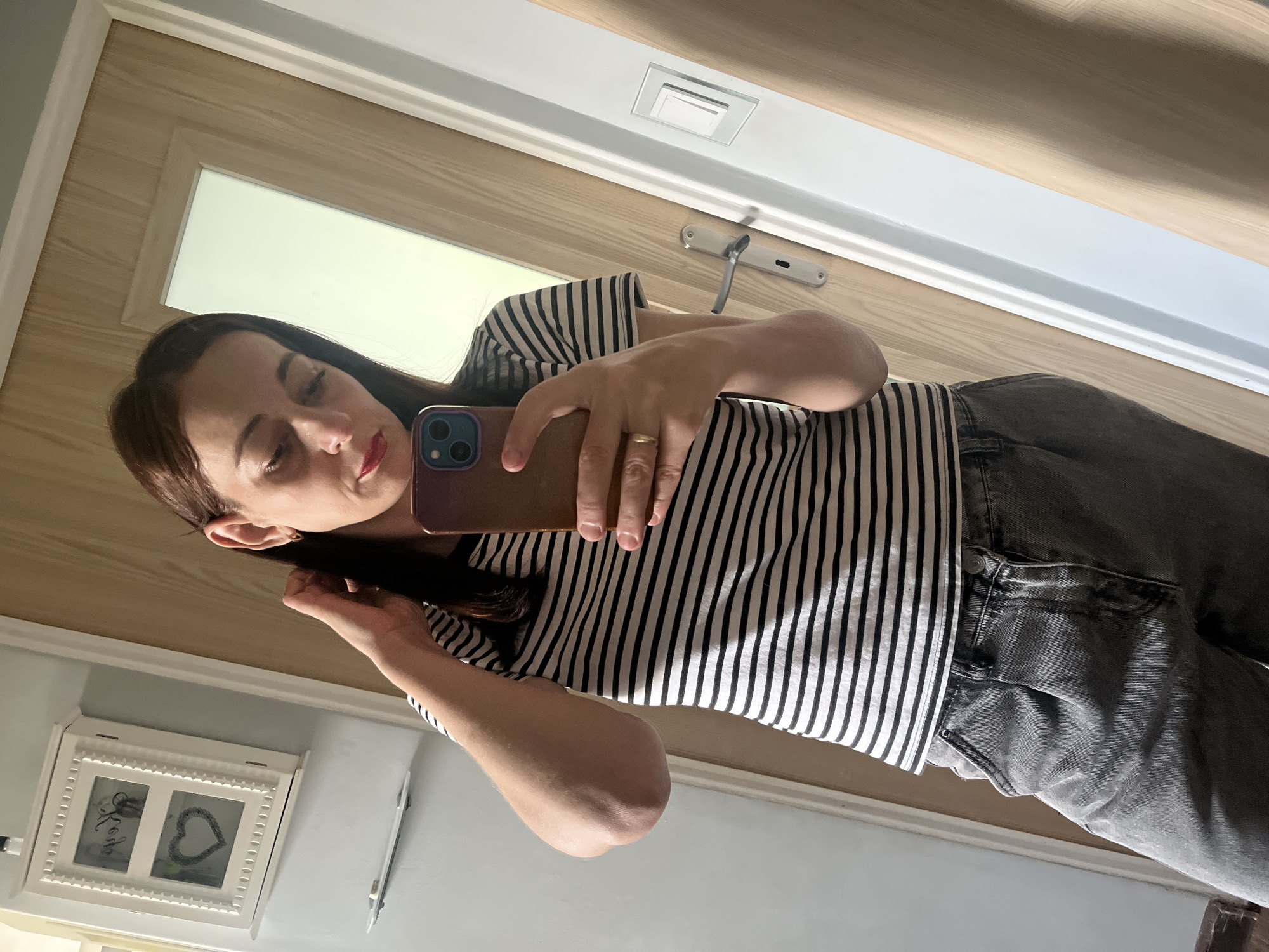 A woman is captured in a mirror selfie, standing indoors. She has dark hair and is wearing a black and white striped short-sleeved top and grey jeans. Her lips are adorned with red lipstick. She holds a smartphone with a reddish-brown case, featuring multiple camera lenses, in front of her to take the photo, looking towards the screen. A gold-colored ring is visible on her right hand.
The background primarily features a light-colored wooden door with a rectangular translucent panel and a metallic silver door handle and lock mechanism. Above the door handle, a square light switch or similar fixture is mounted on the wall. To the lower left of the woman, a white, ornate picture frame with two compartments is visible. One compartment appears to have the word "love" written in it, and the other contains a heart shape. The lighting is bright, suggesting it is daytime.