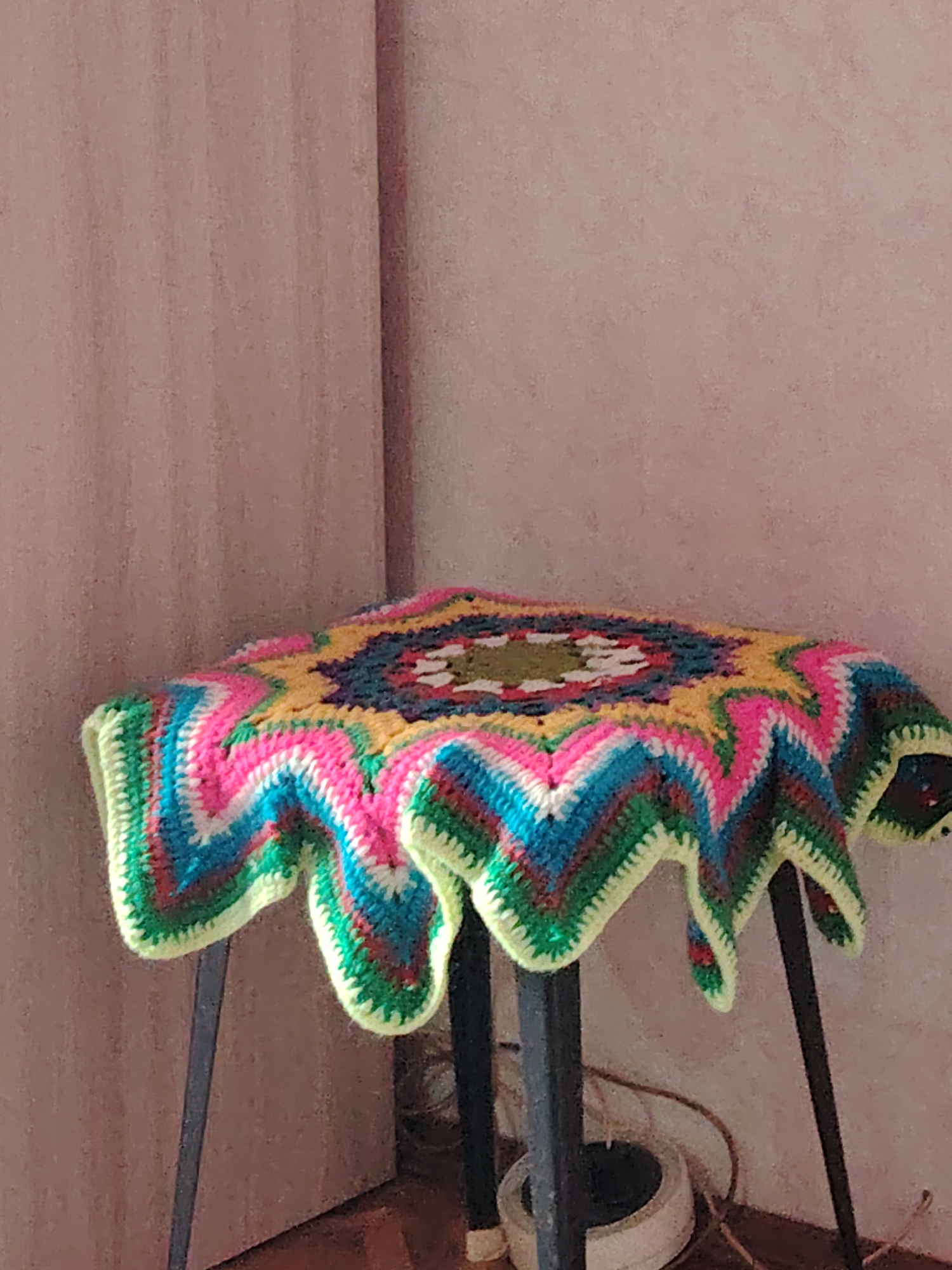 A vibrant, star-shaped crocheted blanket or mat is draped over a dark-legged stool or small table, positioned in the corner of a room in Yanaul, Russia. The crocheted item is a riot of color, featuring concentric patterns in bright pink, yellow, teal, blue, white, green, and a deeper shade like maroon or dark green. It has a distinctive zigzag or chevron design radiating outwards from the center, culminating in a wavy, scalloped edge bordered in bright lime green.
The stool or table supporting the blanket has slender, dark-colored legs, likely metal, with three visible in the frame. The background consists of two walls forming a corner. The wall on the left and center has a light pinkish-brown or mauve hue with a subtle vertical striation texture. The wall on the right appears to be a slightly different shade or texture of the same color, creating a distinct corner. The floor in the lower part of the image is a darker brown, possibly wood or carpet. On the floor near the stool's legs, a white circular object, which appears to be a base or electrical component, is visible, along with some thin cables or wires.
The scene is an indoor, still life arrangement, with no activity taking place. The lighting is ambient, without strong shadows to indicate a specific time of day. No people are present, and no text is visible in the image. The overall impression is one of handmade craft and a cozy indoor setting.