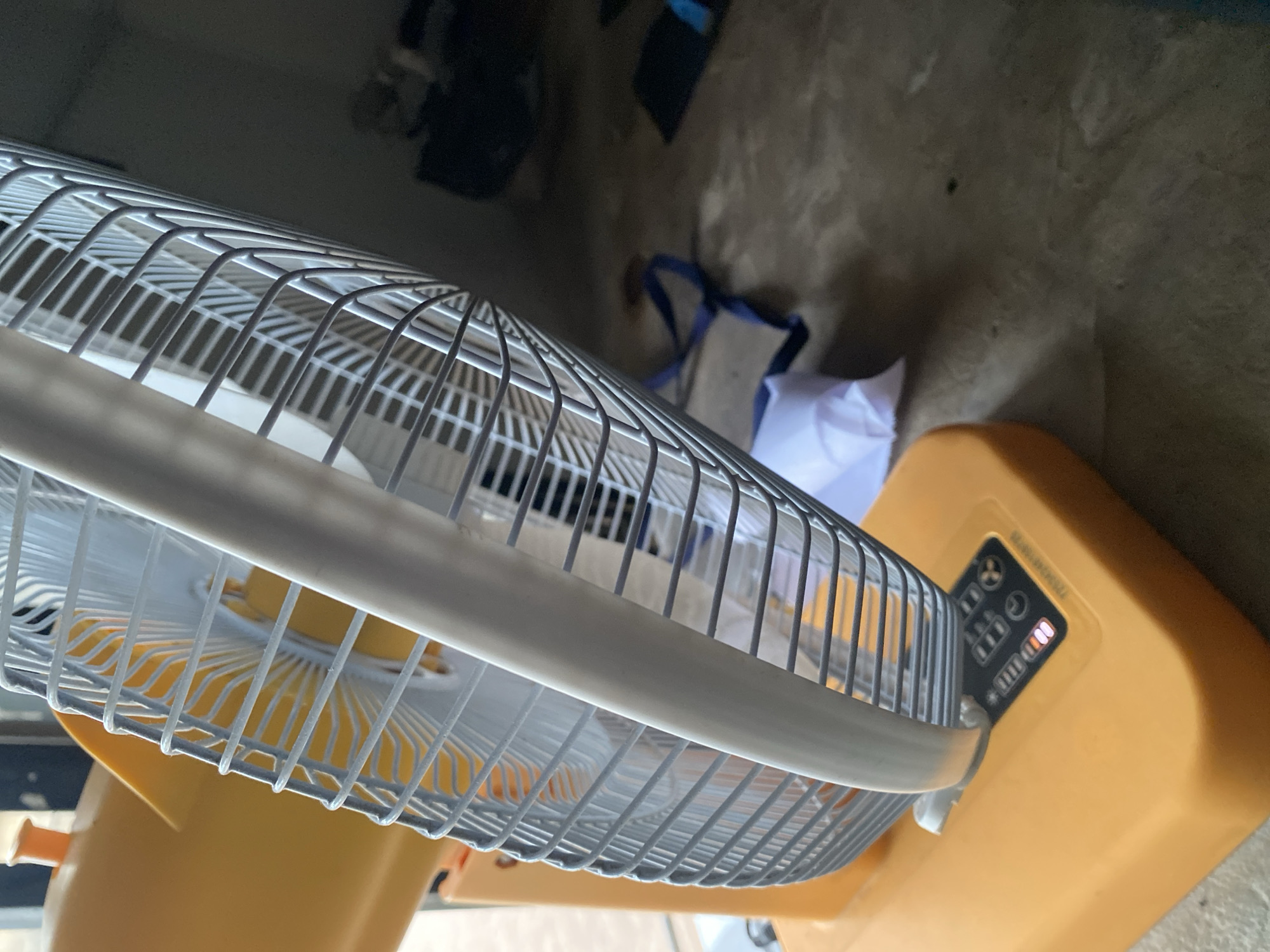 An outdoor, close-up shot shows a yellow electric fan. The fan is angled, with the front grille dominating the foreground. The grille is silver and made of thin metal bars arranged in a circular pattern. A white plastic handle or frame runs around the edge of the grille. The fan's base is a bright yellow, square shape, and a small control panel with lights is visible on the front of the base. The background is out of focus and appears to be an indoor space with a concrete floor. There is a blue bag and some white fabric on the floor. The lighting is dim. This image was taken in Jalingo, Nigeria.