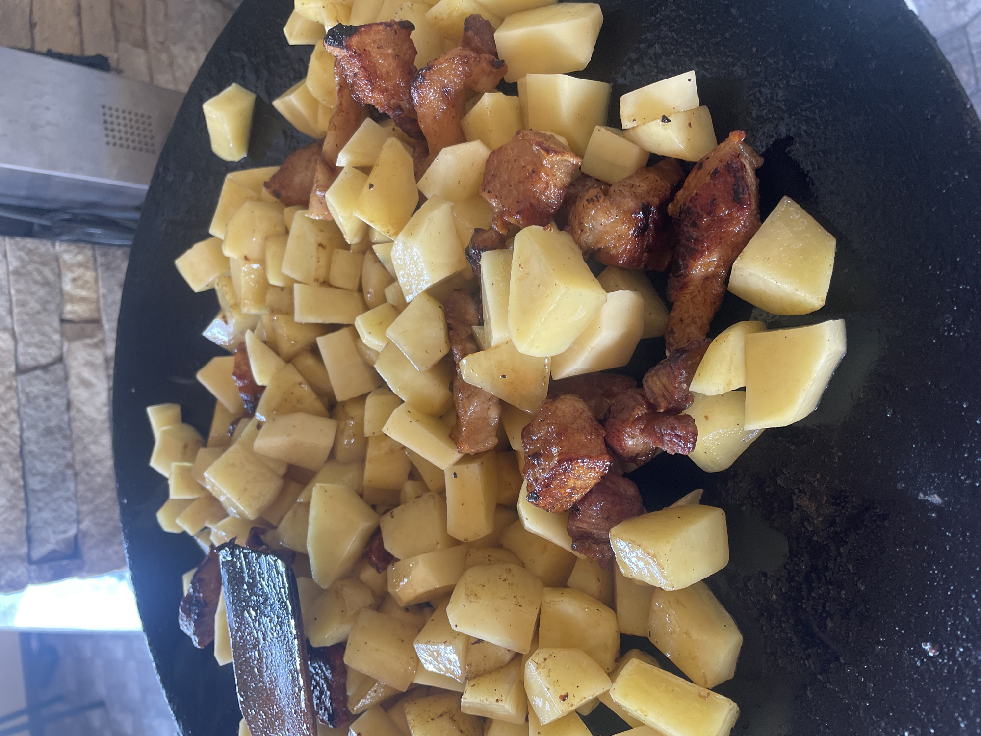 The image displays a close-up, overhead perspective of a large, dark, circular metal griddle or pan containing food actively cooking. Numerous irregularly diced potatoes, predominantly light yellow to off-white, are spread across the surface, mixed with browned pieces of meat, consistent with pork or chicken, exhibiting caramelization and dark seared edges. A dark wooden cooking utensil, possibly a spatula, is visible on the left side of the griddle, resting among the food. The background on the right features a dry-stacked stone wall constructed from rectangular, textured blocks in shades of grey and beige. Adjacent to this wall, a dark, vertical metallic element with a grid-like perforated pattern is partially visible. The upper-left background is out of focus, revealing ambient light and indistinct structural elements. The scene depicts the preparation of a meal in an outdoor or semi-outdoor environment located in Petrivka, Ukraine. No individuals are present in the frame.