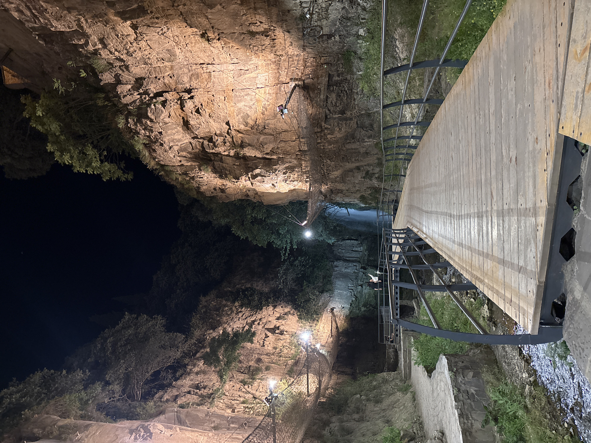 A wooden boardwalk with a dark metal railing curves into the foreground on the right, leading towards a narrow gorge. On the left, a stone path winds along the base of a steep, rocky cliff face. Suspended across the gorge at different heights are mesh net bridges, illuminated by several bright lights. Trees and vegetation grow from the rocky cliffs. In the distance, a waterfall is visible, with two people standing on a bridge near its base. The scene is illuminated by artificial lights, indicating it is nighttime or a very dark environment.