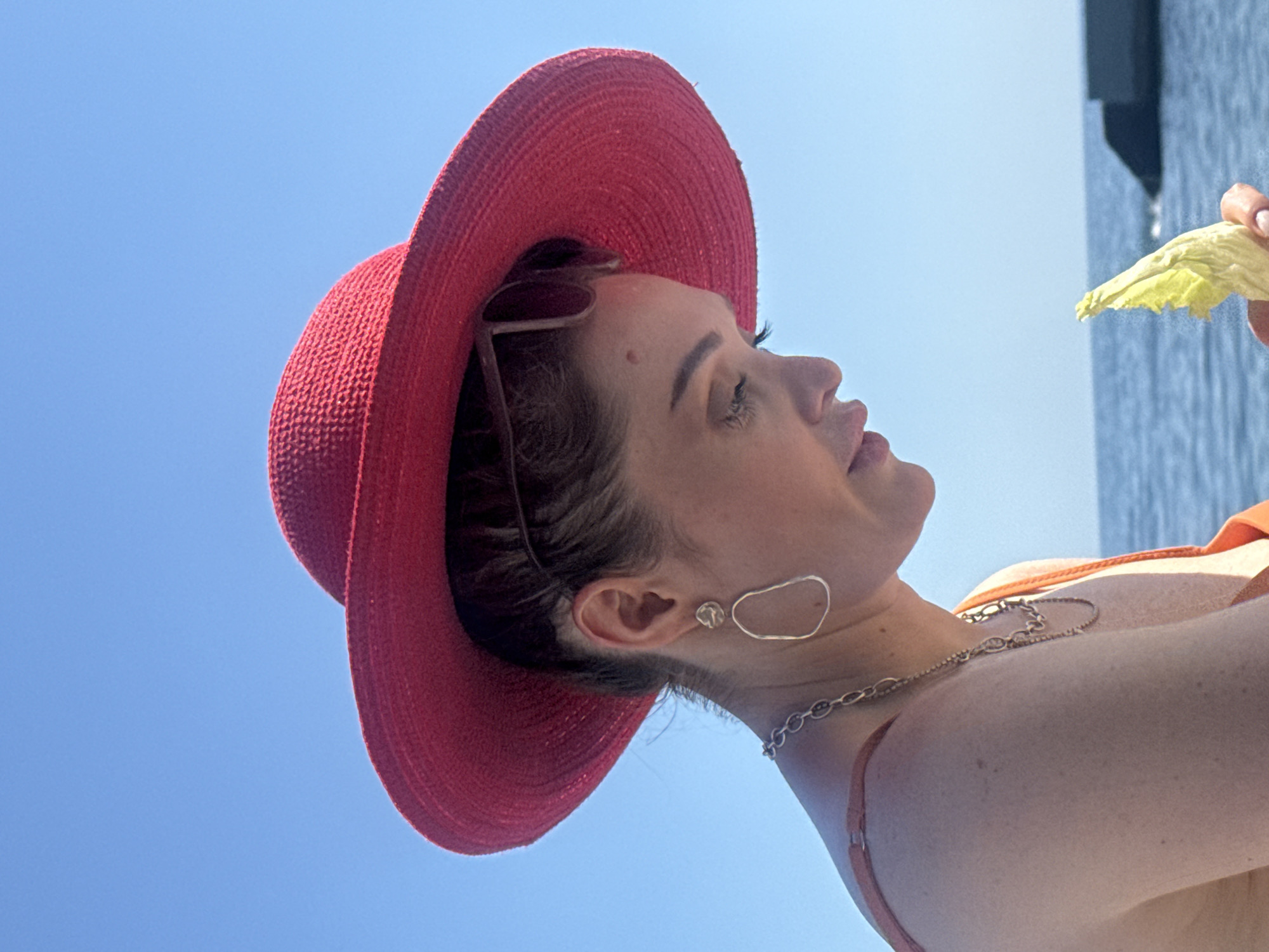 A young woman wearing a wide-brimmed red hat and an orange top is pictured from the chest up, looking down at a piece of lettuce in her hand. She has on a silver necklace and abstract, elongated earrings. Her sunglasses are perched on top of her hat. The background is a clear blue sky and the ocean, with a dark, man-made structure visible in the distance on the right. The image appears to have been taken outdoors on a sunny day, likely in a coastal location. The woman is the sole subject in the foreground.