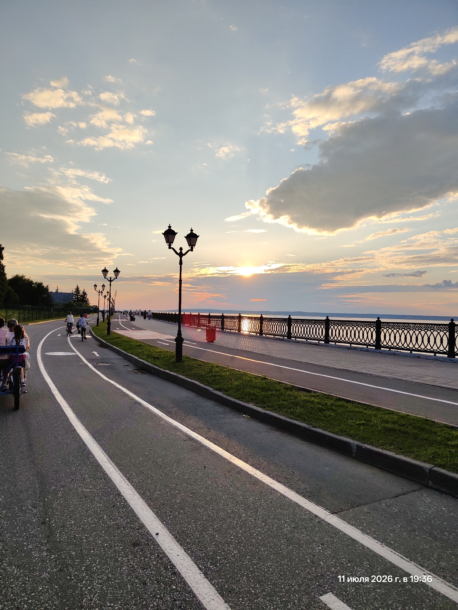 The image depicts a waterfront promenade at sunset. On the left, a paved road with white lane markings curves away from the viewer. A multi-person bicycle, carrying at least three people, is moving away on this road. Further down the road, several individuals are riding bicycles. The promenade itself is to the right of the road, separated by a strip of grass. It features decorative lampposts and a black metal railing overlooking a body of water. A red bin is visible on the promenade. The sky is partly cloudy with the sun setting over the water, casting a warm glow. The timestamp "11 июля 2026 г. в 19:36" is visible in the lower right corner.
