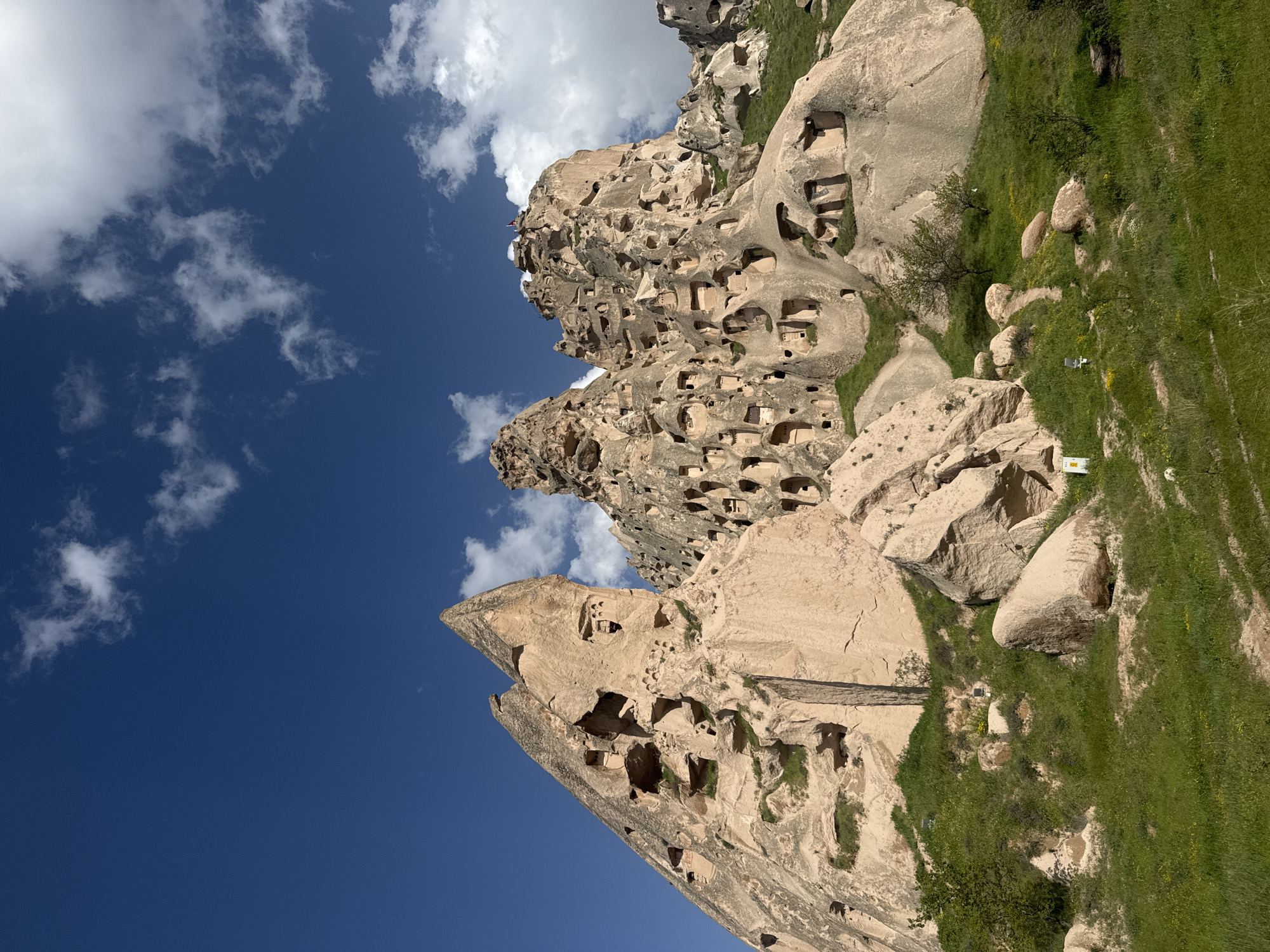 A vertical outdoor shot depicts Uçhisar Beldesi, Turkey, featuring a landscape dominated by distinctive rock formations with numerous carved-out openings and caves, suggesting ancient dwellings or storage spaces. The sky, a deep blue, is speckled with scattered white clouds, contrasting with the beige and tan tones of the rocks. Lush green grass grows at the base of the rock formations, offering a contrast in color and texture. A small white object rests on the grass among the rocks. The composition is tilted, with the rock formations extending diagonally across the frame from lower-left to upper-right.