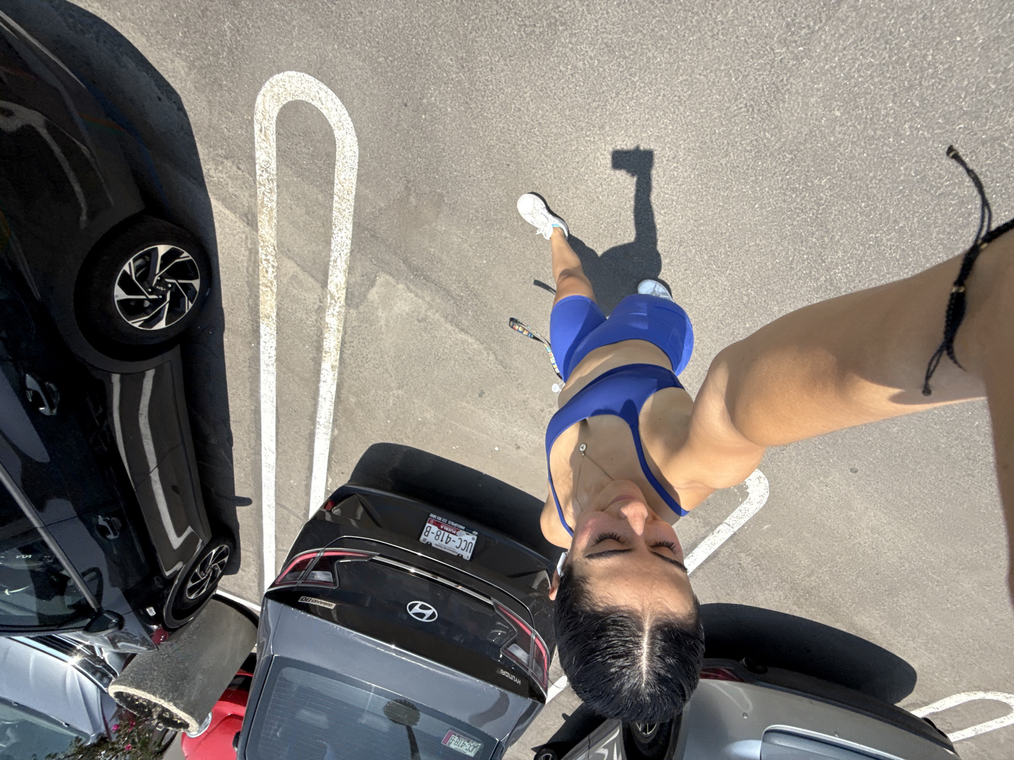 The image depicts a woman standing in a parking lot in Puebla, Mexico. She is wearing a blue sports bra and shorts, white sneakers and has a black bracelet on her right wrist. Her arm is extended in front of her, likely holding the camera. Her head is tilted towards the ground. The parking lot contains several cars parked in marked spaces. A black car is on the left, and a grey Hyundai with a license plate "UCC-418-B" is near the center. Parking space markings are visible on the asphalt. The overall lighting indicates it is daytime with a clear sky.