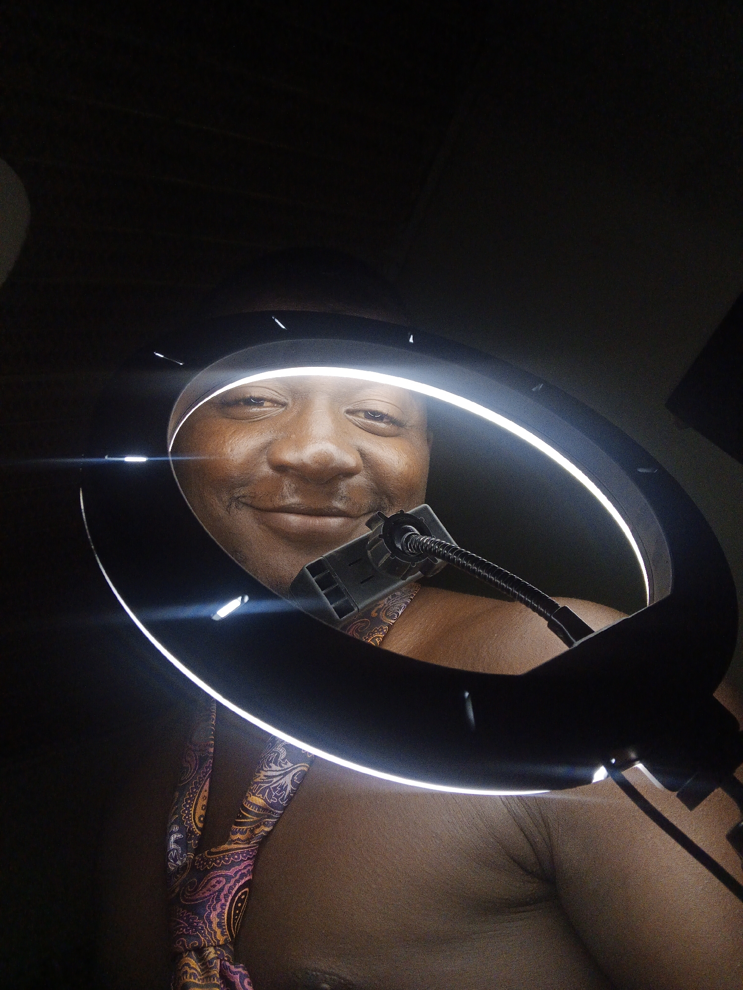 A person is shown in a close-up selfie, with a ring light illuminating their face and upper body. The person has dark skin and is smiling. They are wearing a colorful patterned tie around their neck. The background is dark and indistinct, suggesting an indoor setting. The image appears to be taken in a location in Akwanga, Nigeria.