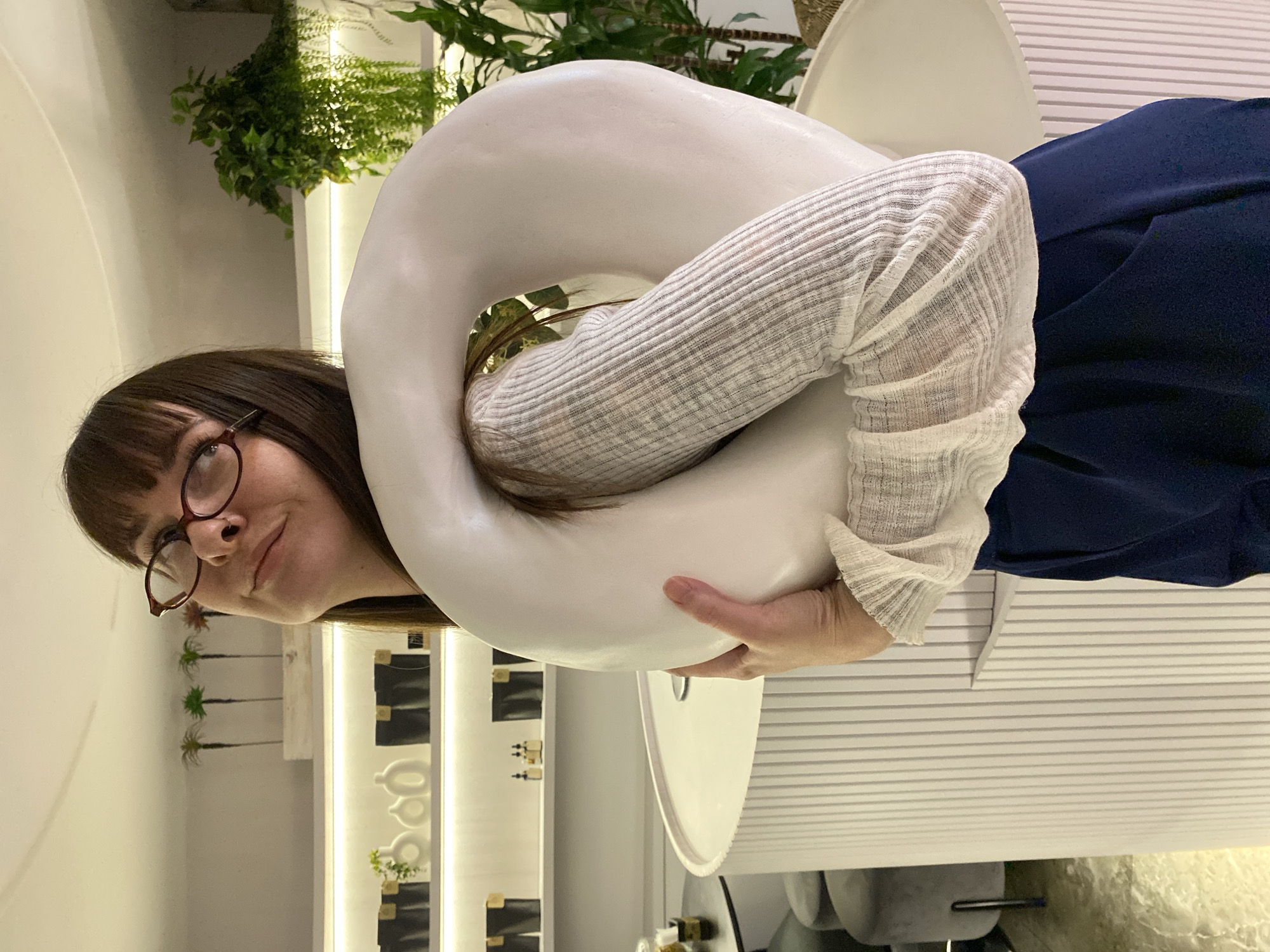 An eye-level portrait shows a woman holding a large, smooth, white, C-shaped object, possibly a sculptural piece, in a modern indoor setting in Votkinsk, Russia. The woman, with dark brown hair and bangs, wears round, reddish-brown framed glasses and a light-colored, ribbed long-sleeved top. Her left arm is threaded through the object, and her right hand supports it near her waist. She smiles gently, looking towards the viewer.
The background reveals a brightly lit interior with a clean, contemporary aesthetic. Lush green foliage, possibly ferns, hangs from above on the left side. To the right and behind the woman, there's a white wall panel with horizontal grooves. Further back, a display area features shelves with small bottles or containers and abstract white decorative objects, including small palm tree figurines. The overall ambiance is chic and well-lit, suggesting a boutique, gallery, or modern living space. No text is visible in the image.