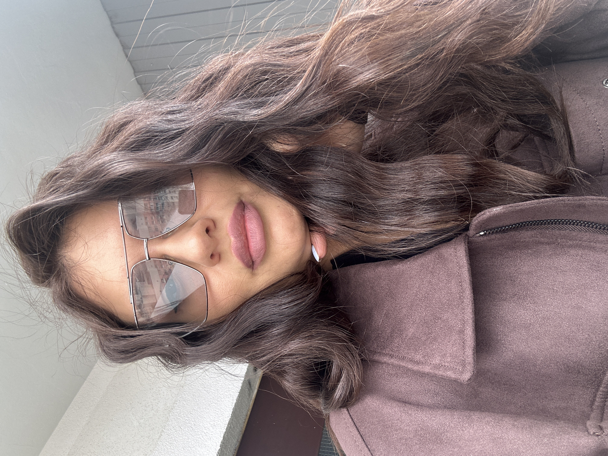 A young woman with long, wavy brown hair is wearing glasses and a brown coat. The location is potentially outside, indicated by the bright, overcast sky. Given the description of the prompt, one potential location is the city of Khimki, Russia.