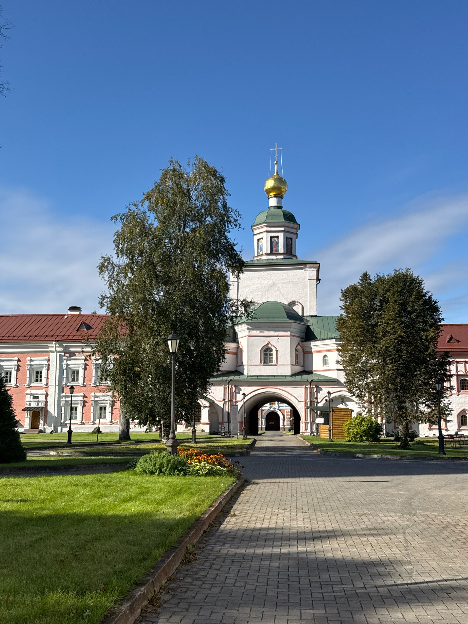 This is a forward-facing, eye-level, medium shot of the Valday Iversky Monastery in Valday, Novgorod Oblast, Russia. The image captures the monastery's main gate, with a tall, white bell tower topped by a golden dome and cross in the center. To the left and right of the gate are the monastery's red-roofed buildings, featuring traditional Russian architecture with white trim and arched windows. A paved walkway leads directly to the gate, bordered by a well-maintained green lawn on the left and flowerbeds. Several large trees, including a prominent birch tree, are positioned in front of the buildings, and a lamppost stands near the walkway. The sky is clear and blue with some wispy clouds. The scene appears peaceful and serene, with no people visible.