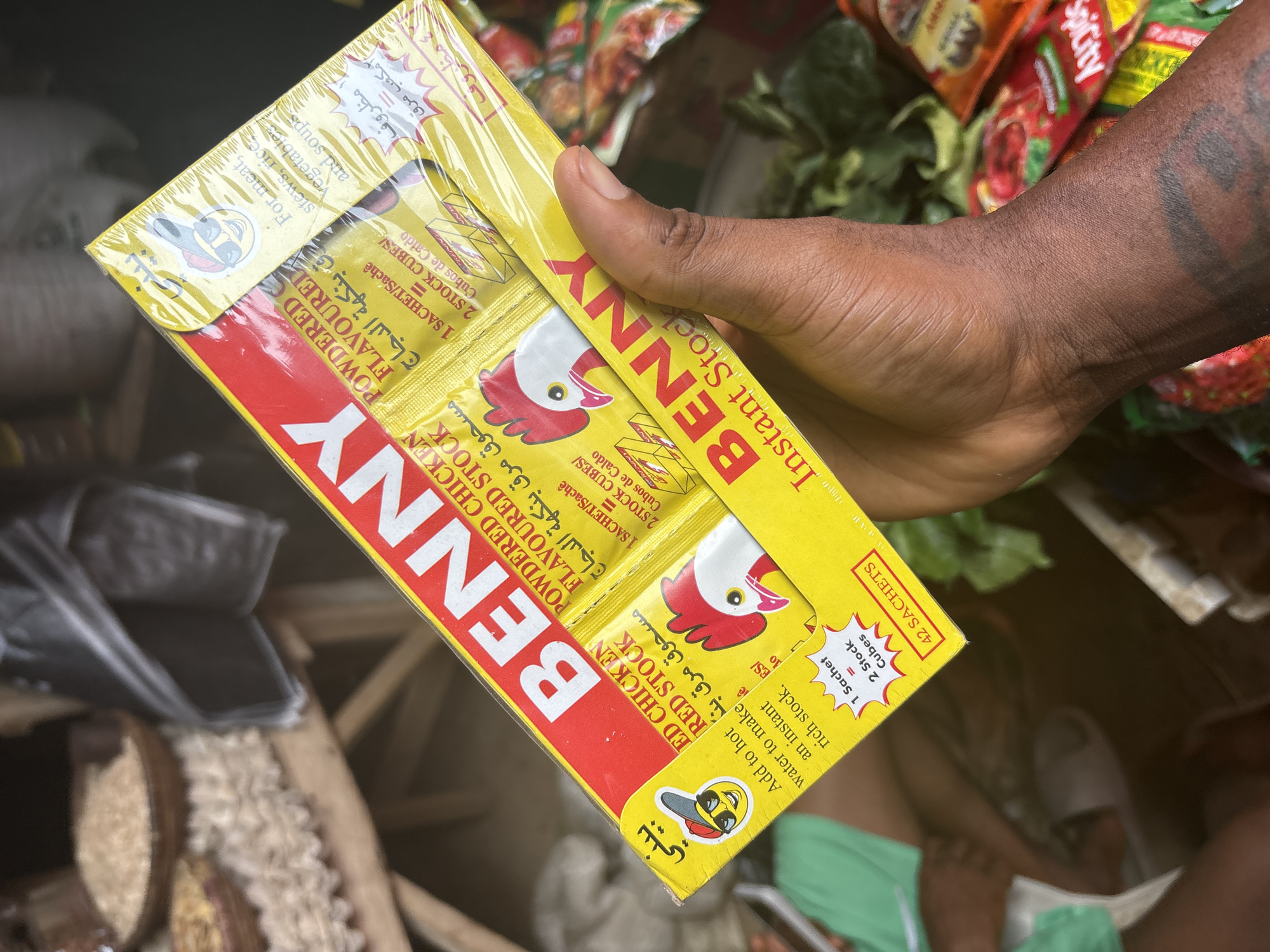 An eye-level shot captures a dark-skinned hand holding a brightly colored yellow and red packet of "BENNY Instant Stock" in an indoor market setting, likely in Iddo Tudunwada, Nigeria. The hand, adorned with visible tattoos on the forearm, presents the product vertically.
The packaging prominently displays "BENNY" in bold red capital letters. It is labeled as "POWDERED CHICKEN FLAVOURED STOCK" and features a cartoon chicken head logo. The packet indicates it contains "42 SACHETS" and provides instructions to "Add to hot water to make an instant rich stock," recommending its use "For meat, stews, rice, vegetables and soups." A conversion graphic states "1 Sachet = 2 Stock Cubes." The text on the packaging is in English, Arabic, and possibly other languages.
The background is blurred, revealing glimpses of other market goods, including various packaged items, one labeled "Spicity," and fresh green leafy vegetables, suggesting a food stall or market aisle. In the lower right, part of a person's leg wearing a light-colored slipper is visible, implying someone is seated or squatting nearby. The scene is illuminated by ambient indoor lighting, highlighting the product.