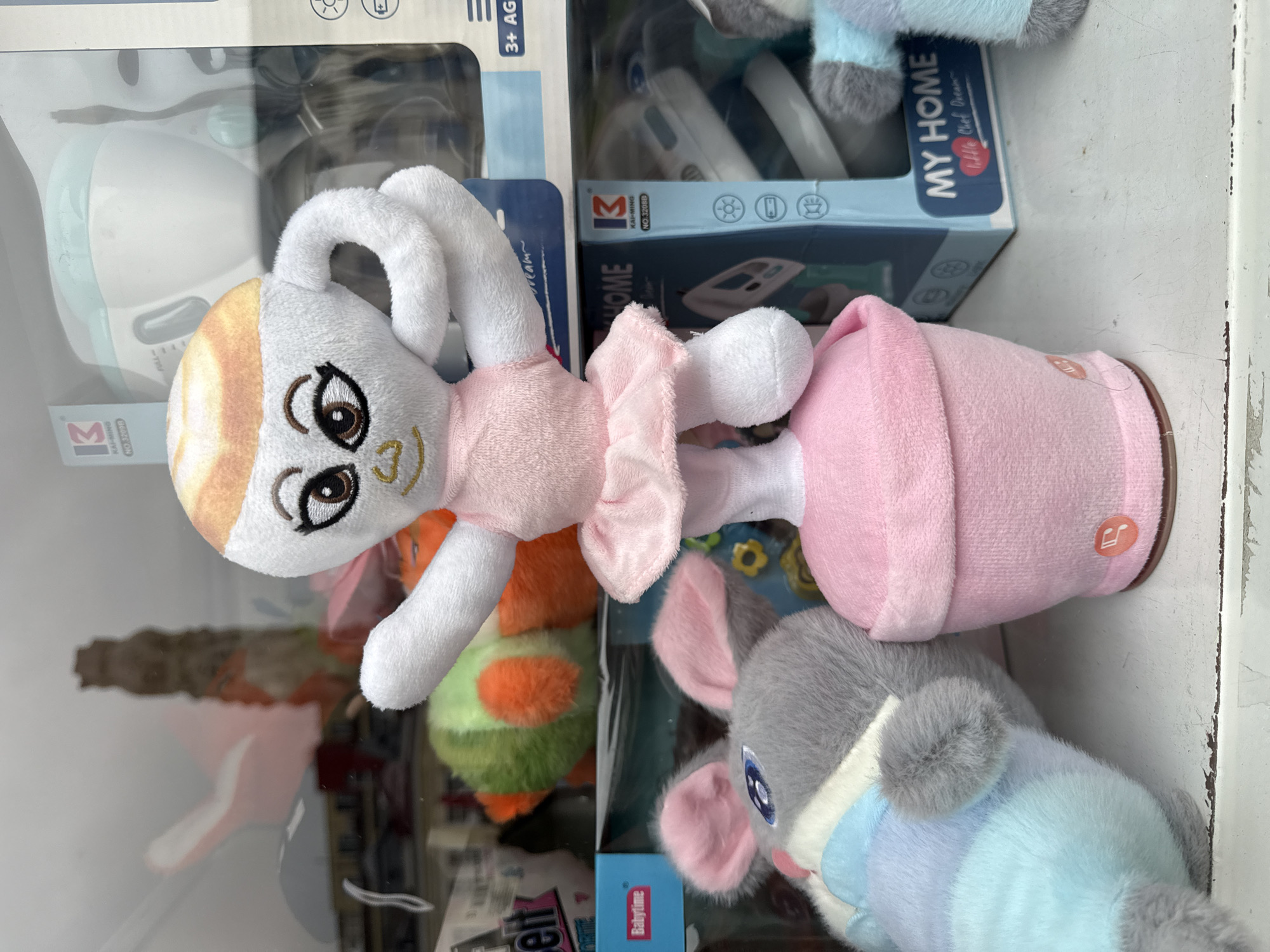 This media file presents a vertical close-up view of plush toys and toy product packaging, likely situated on a shelf or display in a retail environment.
The main subject is a white plush toy, resembling a rabbit or bunny, wearing a light pink ballet-style outfit including a leotard and a ruffled skirt. It features large, expressive brown eyes with prominent eyelashes and a gentle smile. The top of its head has a distinct orange-tan gradient coloring. The plush is positioned within a soft pink cylindrical container or base, which bears a small orange circular tag embossed with a music note icon.
Below and around the central toy, other plush toys are partially visible. To the left, a green and orange plush item is seen, while to the lower right, a gray and light blue plush toy with pink inner ears, possibly depicting a mouse or elephant, is present.
In the background, several toy boxes are stacked, predominantly in white and light blue tones, featuring clear plastic windows that reveal contents resembling small white toy appliances, possibly an iron or similar household item. Visible text on these boxes includes the brand "KAI-MING" with model numbers "NO. 32098" and "NO.32088," "MY HOME," and "Babytime." One box clearly states "liffle Chef Dream" (likely "Little Chef Dream") and specifies "3+ AG," indicating an age recommendation of 3 years and above. Icons on the packaging suggest features like light emission, battery operation, and a temperature or heat function.
The scene is well-lit, characteristic of an indoor commercial space. No people are visible, and the toys are simply on display. The image does not contain any specific visual cues to confirm the location as Shumen, Bulgaria, beyond the context provided.