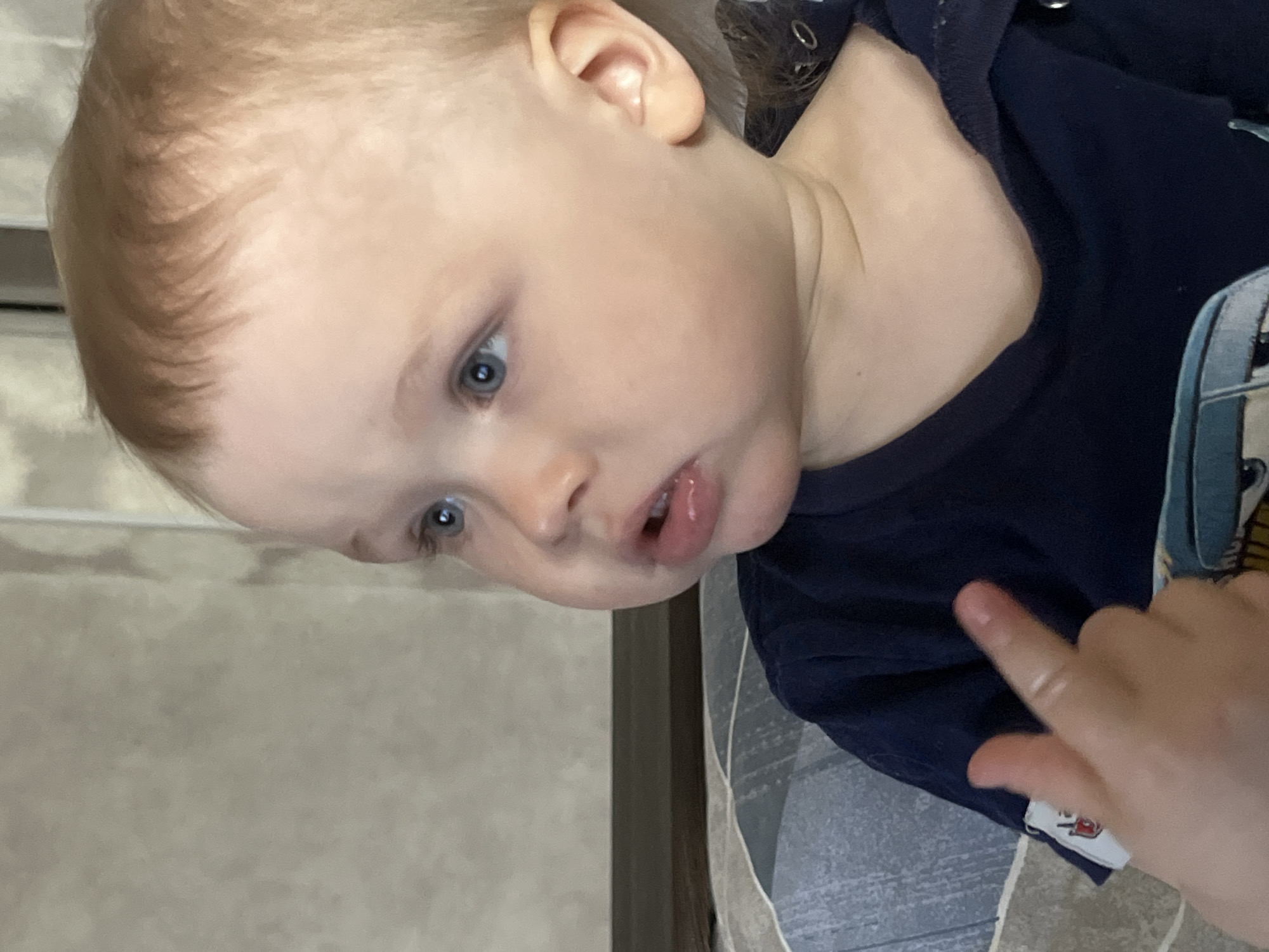 A fair-skinned child with blue eyes and light blonde hair is visible from the ear up to the top of their head. They are wearing a navy blue garment. Part of a hand with an extended index finger is in the lower right corner of the image. The background is a light, neutral tone. It is not possible to determine if the image was taken in the city of Chaikovsky, Russia.