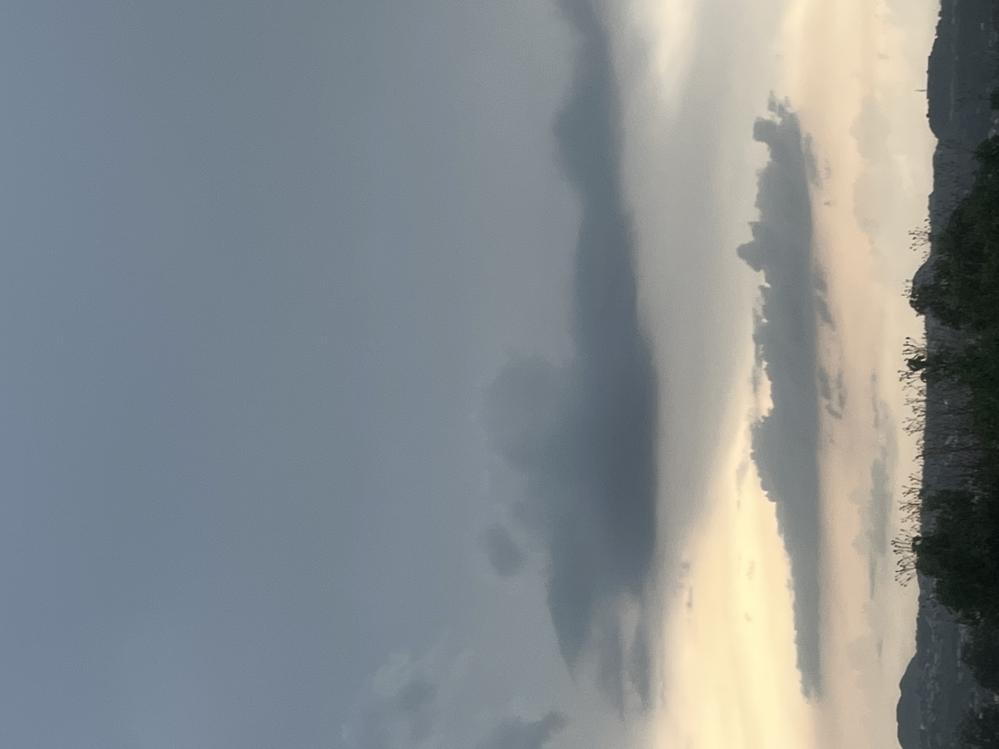 The image shows a cloudy sky over a landscape that is not identifiable as Gura Top, Nigeria. The sky is filled with various shades of gray clouds, with some lighter, warmer tones near the horizon, suggesting either sunrise or sunset. In the foreground, silhouetted trees and vegetation are visible against the horizon. Distant hills or mountains can be seen, with hints of buildings or structures on them. The overall atmosphere is overcast and muted.