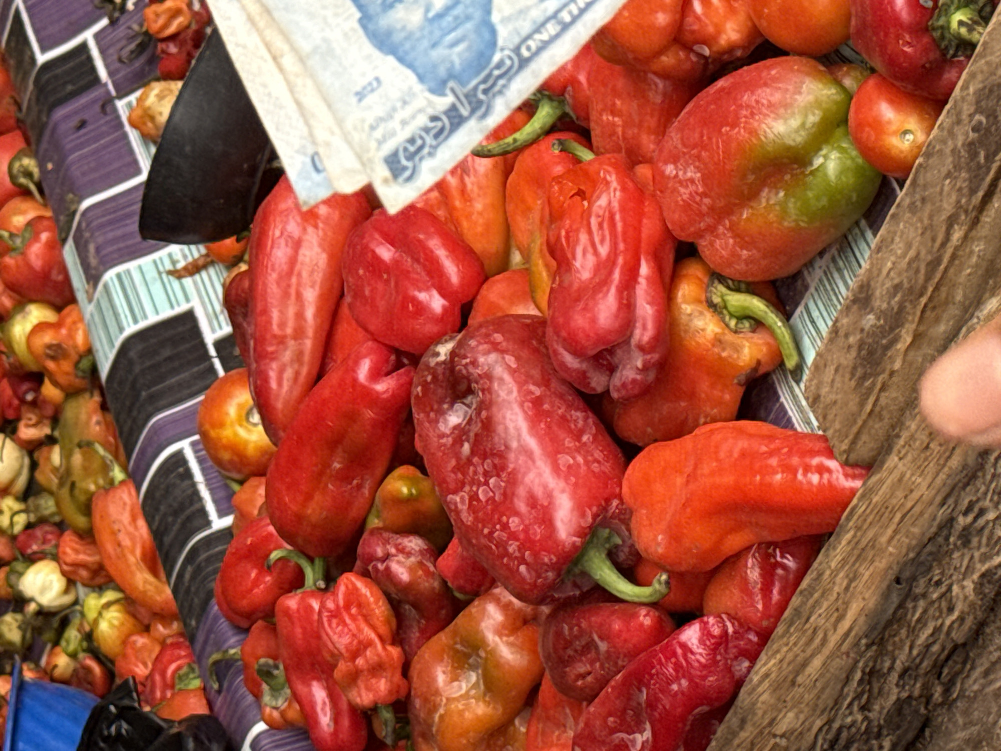 A close-up captures a vivid array of red peppers, likely displayed for sale. The peppers fill a section of a wooden crate or stall. A bill with Arabic script and a blue portrait is placed on top. The arrangement suggests a market setting. The presence of the Arabic bill indicates the sale may be occurring in a market in Abuja, Nigeria. A large black cleaver lies near the peppers. A tablecloth with a geometric pattern rests to the left of the peppers. Parts of a hand can be seen on the right side of the wooden crate.