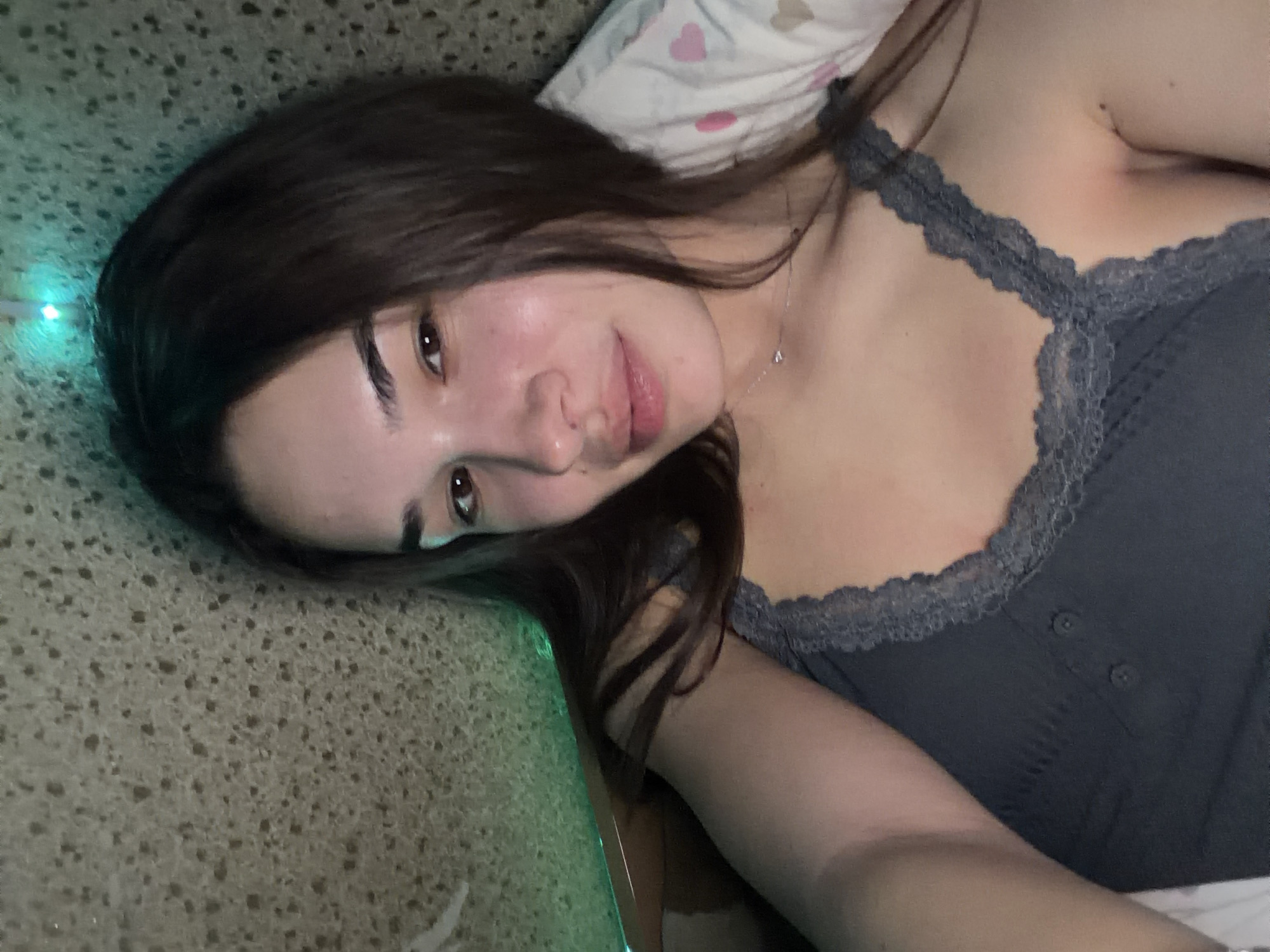 A woman is lying down, possibly in a bedroom, oriented diagonally in the frame. She is wearing a gray tank top with lace trim. She has long brown hair, fair skin, and appears to be looking directly at the camera with a slight smile. A pillow with a pink heart pattern is visible behind her head. The background includes a surface with a speckled texture that could be a countertop or wall and there is a green glow originating from the left edge of the frame. There are no indications of being located in Sofia, Bulgaria.