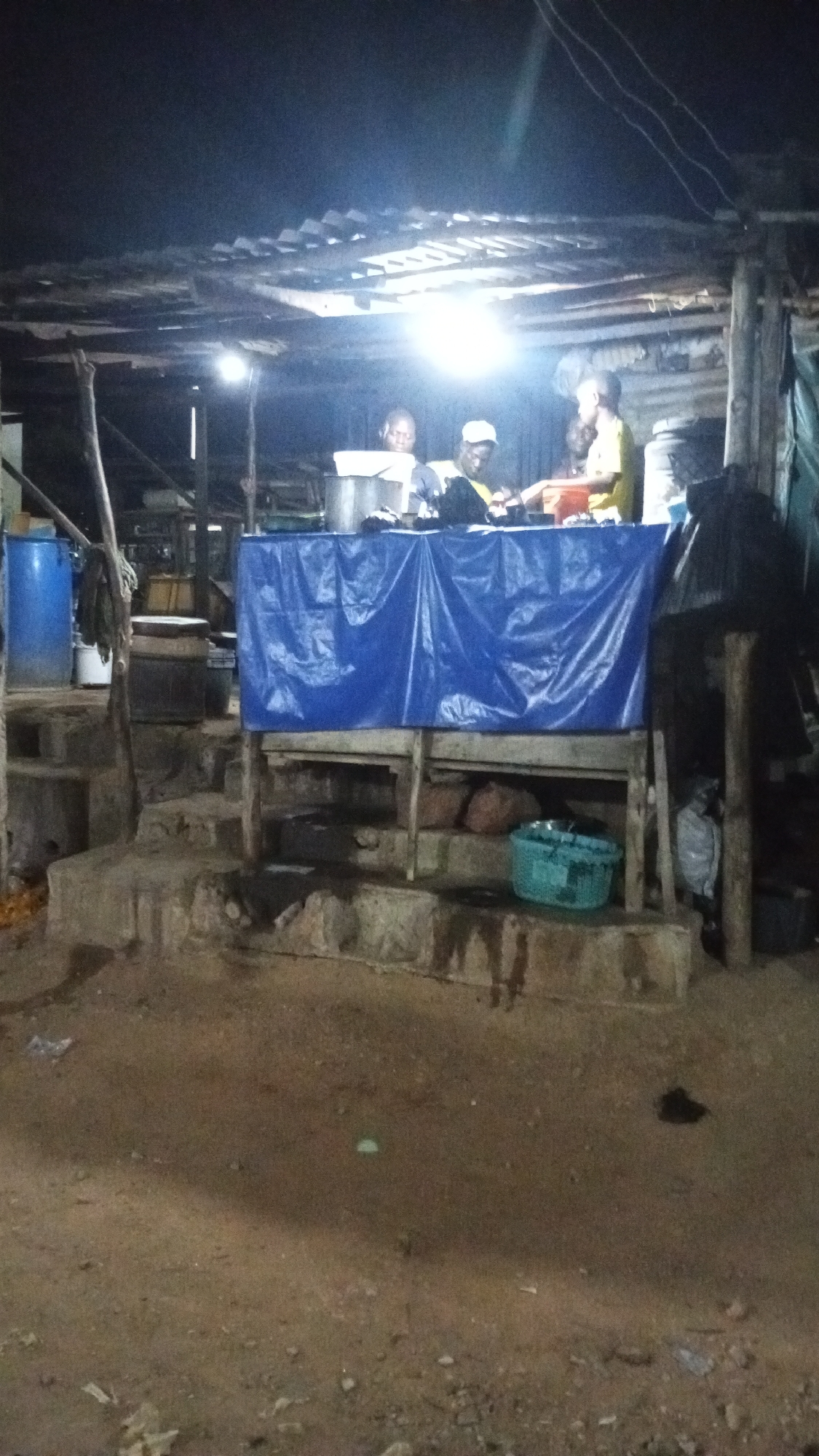 This image depicts a makeshift outdoor food stall in Adankolo, Nigeria, operating under dim artificial lighting. Three individuals are visible behind a table draped with a blue plastic sheet, possibly preparing or serving food. The stall appears to be constructed with corrugated metal roofing and wooden supports. Various containers, including a blue barrel and other vessels, are present. The ground is dirt. There are no street signs, store signs, or indicators of formal business activity visible. The overall atmosphere is informal and utilitarian.