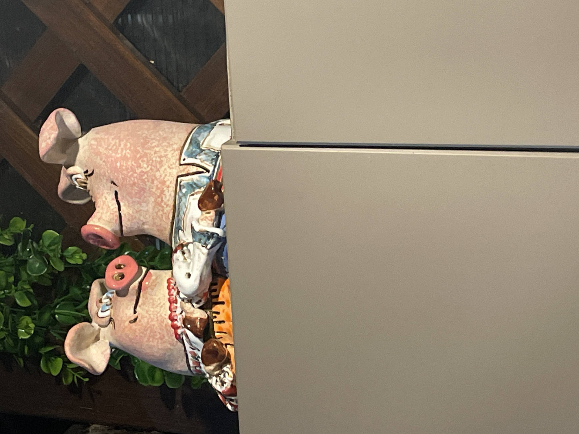 A pair of ceramic pig figurines are perched on top of a gray cabinet. The figurines are pink with black eyes and rosy cheeks. They are wearing colorful, patterned clothing. The figurine on the left wears a red beaded necklace, while the figurine on the right has blue and white striped clothing. Behind the figurines, green foliage is visible, as well as a wooden lattice structure. The cabinet appears to be in a domestic setting.