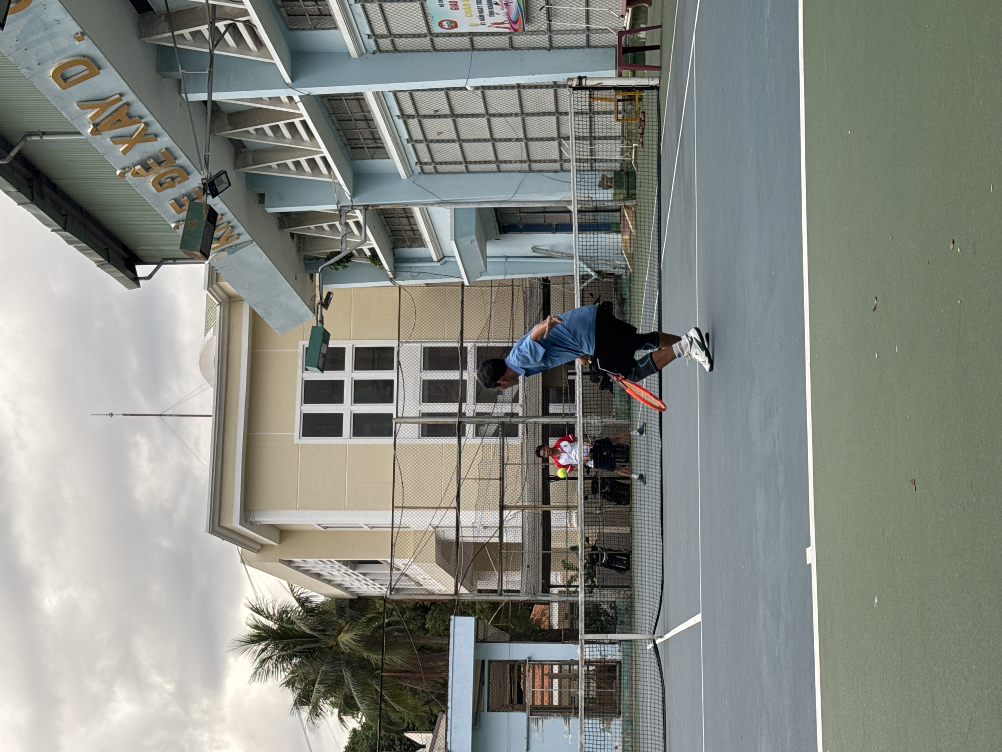 The image depicts a tennis court in Nha Trang, Vietnam, with two men playing tennis. In the foreground, a man in a blue shirt and black shorts is captured mid-serve, his body coiled and racket poised. He wears white and blue tennis shoes and knee support. In the background, across the net, another man in a white shirt with red trim and dark shorts stands ready, holding a tennis racket, with a yellow tennis ball visible in front of him. The tennis court itself is a two-toned surface, with a darker blue court and a greener out-of-bounds area. A net divides the court. Behind the court, there are buildings visible. The building on the right is light blue with distinctive triangular architectural elements and a large sign with Vietnamese text. The building on the left is cream-colored with prominent window frames. Several palm trees are visible in the background, suggesting a tropical or subtropical environment. The sky is overcast with a dramatic, cloudy expanse. The overall scene captures a moment of athletic activity within an urban setting.