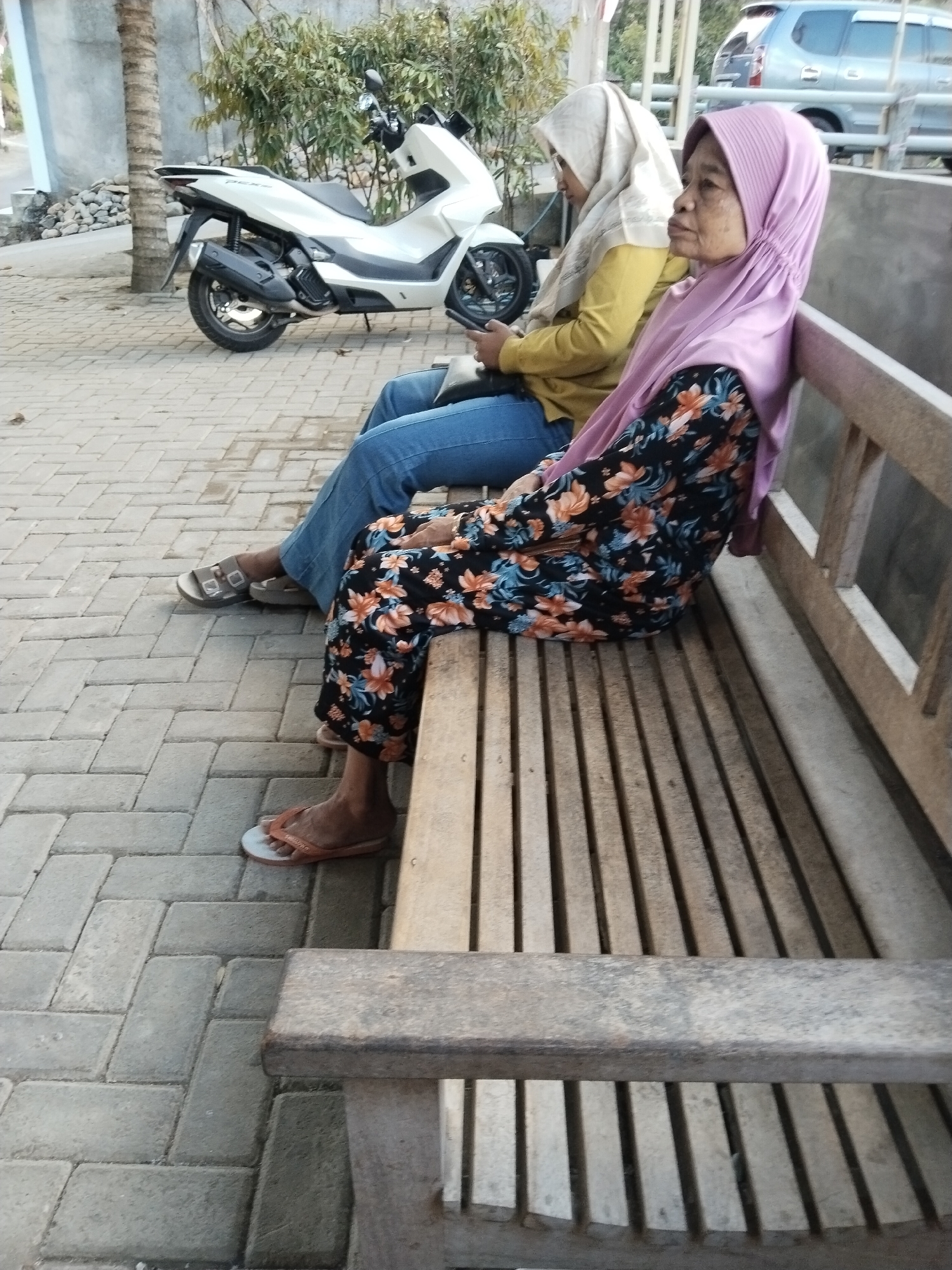 Two women, both wearing headscarves, are sitting on a wooden bench in what appears to be an outdoor public space. The woman on the left is wearing a light-colored headscarf and a yellow long-sleeved shirt, with blue jeans. She is holding a mobile phone in her left hand. The woman on the right is wearing a light purple headscarf and a black long-sleeved dress with an orange and blue floral pattern. She is looking to her right. A white scooter is parked to the left of the bench. The ground is paved with rectangular grey bricks. In the background, there are trees and a light-colored wall. A silver car is visible in the distance. The image is taken during daylight. The scene is consistent with a location in Karanggandu, Indonesia, given the architectural style and vegetation.