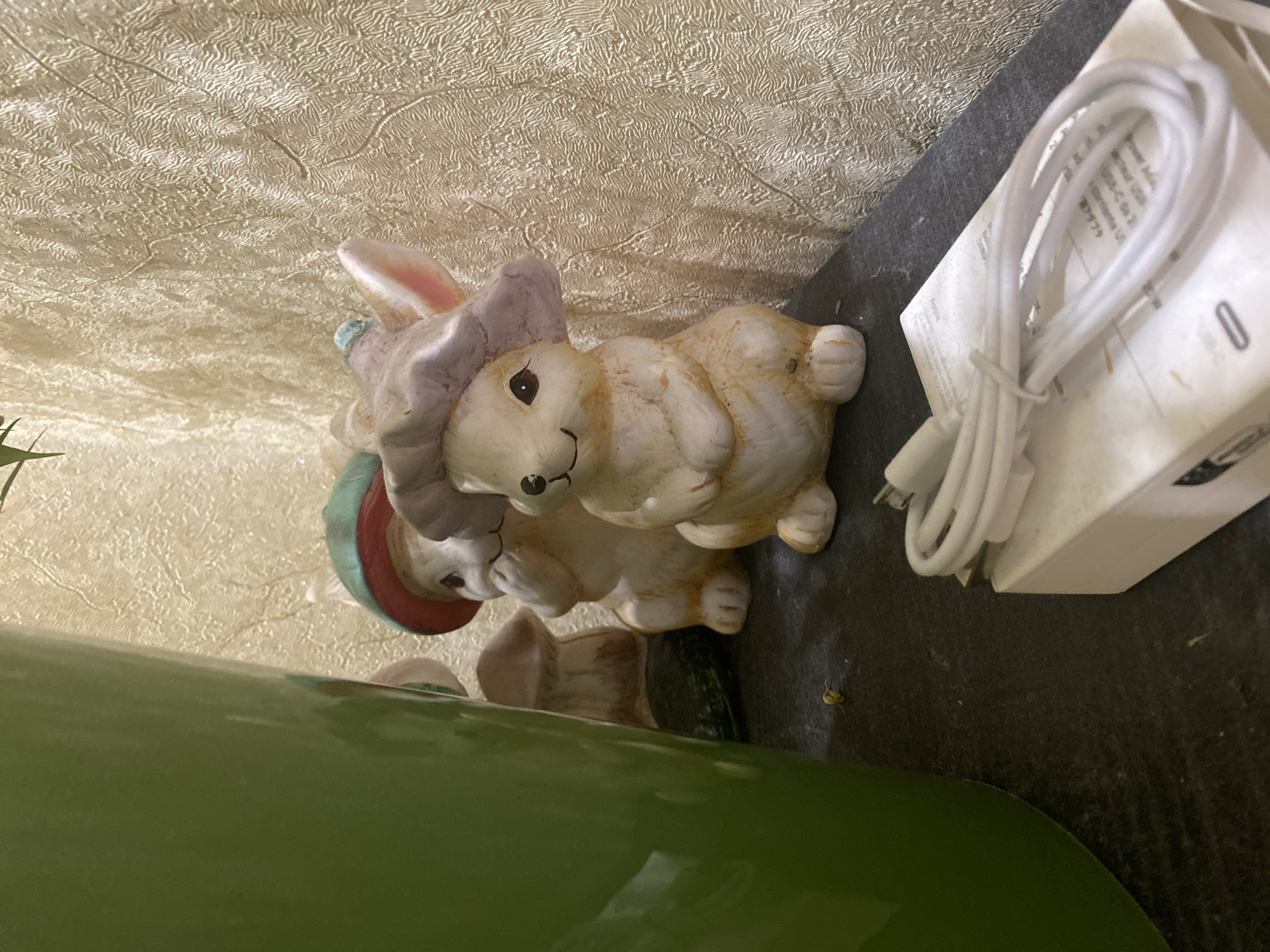 The scene is situated in Tyumen, Russia. The foreground displays two ceramic rabbit figurines positioned on a dark, textured surface. The rabbit in the front-right wears a light lavender hat, features a painted smile, and exhibits a light tan body with areas of brown discoloration. Behind it, partially obscured by a large green object and the first rabbit, another ceramic rabbit wears a green and red hat, with its hands near its face. A brown mushroom-shaped figurine is visible behind this second rabbit. A substantial, smooth, curved green object, likely a plant pot, occupies the left side of the frame. In the bottom-right, a coiled white cable, consistent with a USB-C or Lightning connector type, lies on top of a white power adapter. Visible text on the adapter or its packaging includes "Apple" and Japanese characters. Small yellowish and brownish particles are scattered across the dark surface. The background consists of a light-colored, possibly cream or light gold, textured wallpaper with a distinct ripple or crinkled pattern. The lighting appears localized, creating subtle shadows and highlights. No individuals are present.
