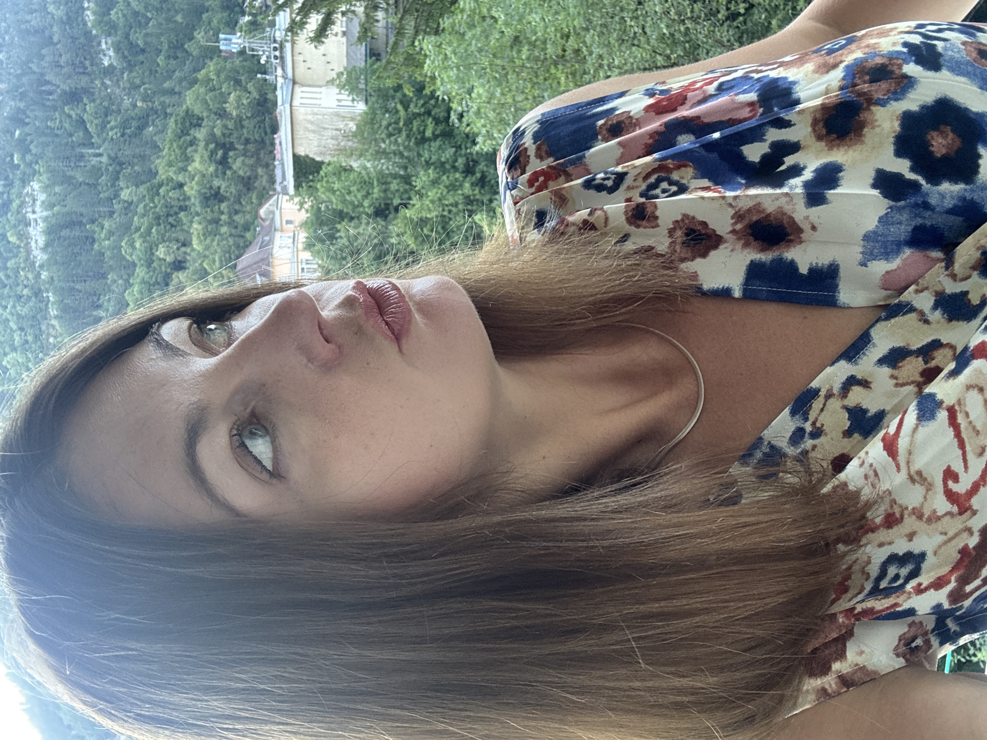 A medium shot shows a woman with shoulder-length brown hair, facing right and looking upward. She has fair skin with freckles and is wearing a sleeveless dress with a floral print of red, blue, and tan. Her lips are slightly pursed, and she has subtle makeup on. A thin silver necklace is visible around her neck. The background is slightly out of focus, showing lush green trees and buildings in the distance, suggesting a mountainous or hilly urban environment. The lighting suggests it is daytime, possibly late afternoon due to the soft glow. The location is identified as Saint-Claude, France.