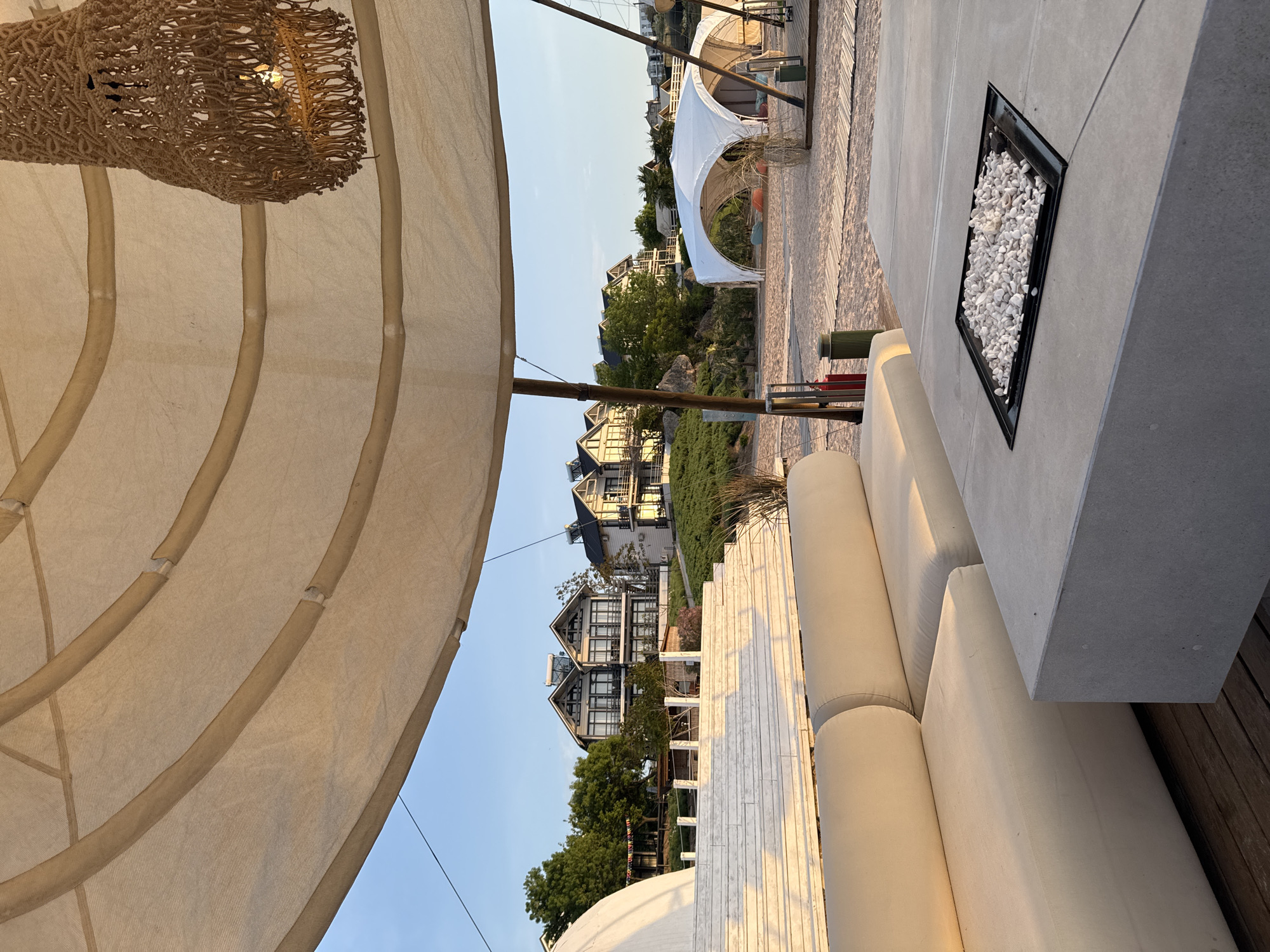 The image captures a scene at an outdoor lounge area, possibly a patio or deck. The location is a cityscape, evidenced by the buildings visible in the background, although the specific city is not identifiable from the image. The image includes a large, tan-colored canvas structure, possibly an awning or a canopy, which provides shade. A decorative wicker light fixture hangs from the structure. A light-colored cushioned outdoor sofa sits next to a concrete countertop, which features a recessed tray filled with small white stones. The foreground is occupied by the awning, the sofa and countertop. The background reveals multiple buildings and vegetation.