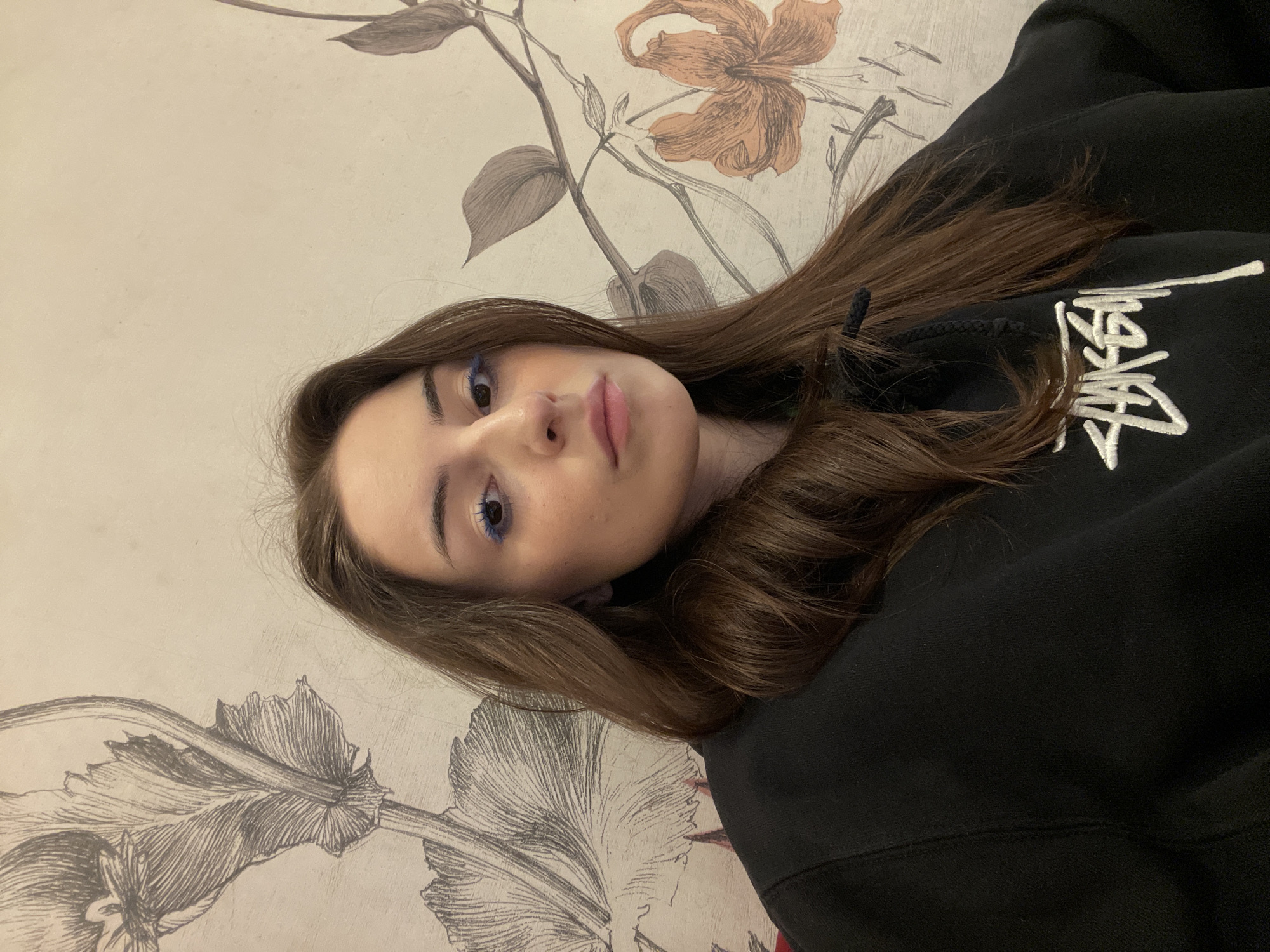 A young woman with long brown hair, wearing blue eyeshadow, is looking at the camera. She is wearing a black hoodie with white text on it. The background is a beige wall with a floral pattern. There is no information to suggest that this image was taken in Moscow, Russia.