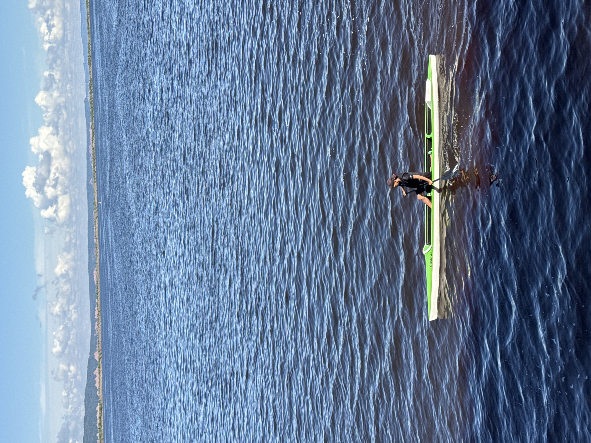 An adult person is seen in a long, narrow, green and white canoe on a dark blue body of water. The person, wearing a dark cap and dark clothing, is seated and holding a paddle, actively paddling the canoe. The canoe has "Albatros" written on its side. The water is rippled, suggesting a light breeze. In the background, a distant shoreline with vegetation and hills is visible under a bright blue sky with scattered white clouds. The scene appears to be an outdoor activity on a calm day.