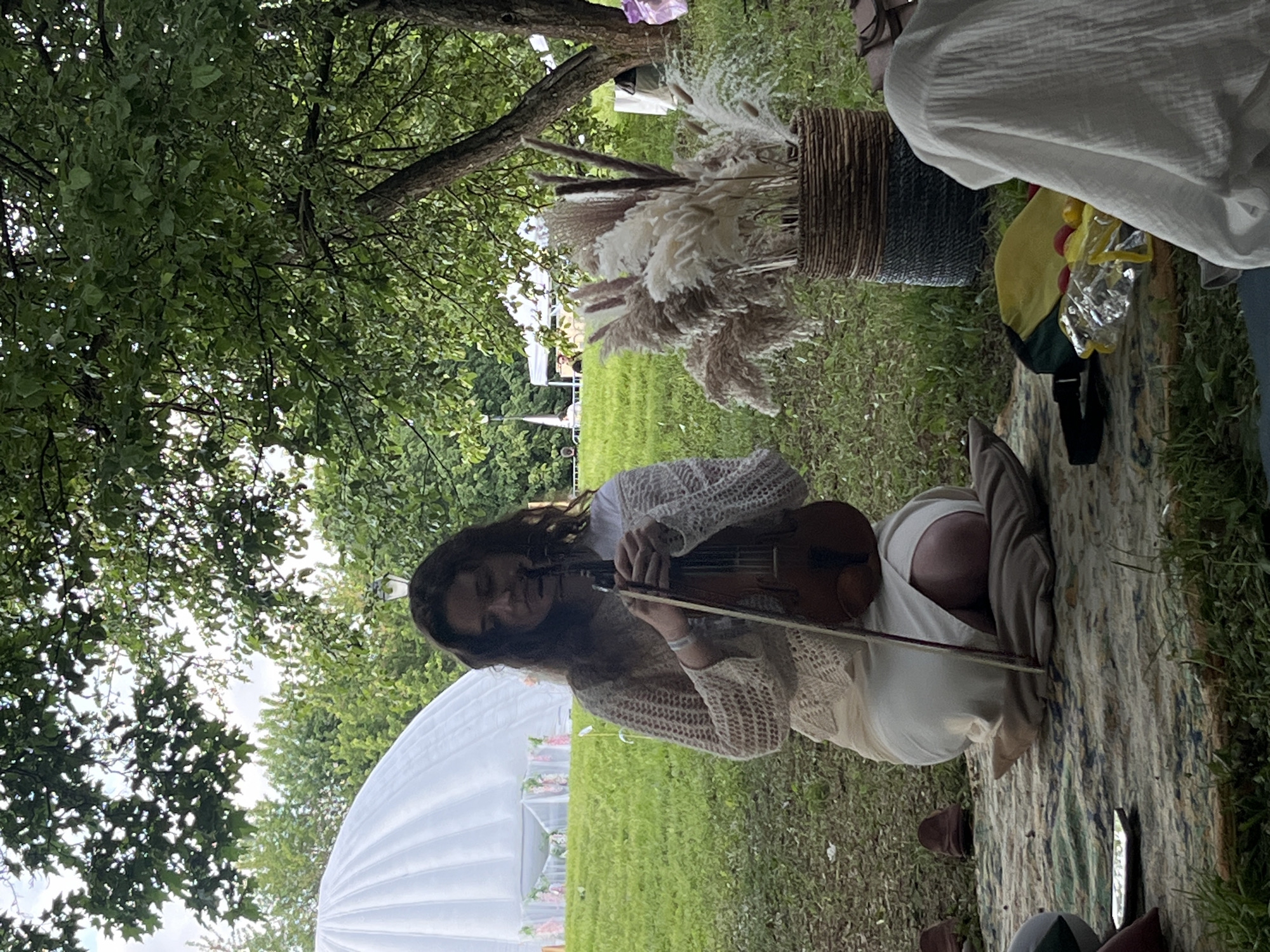 A woman is sitting on a patterned rug on a grassy lawn, playing the violin. She is wearing a light-colored, crocheted top and a white dress or skirt. The scene takes place outdoors, under the shade of trees. To the right of the woman, there's a woven basket filled with decorative plumes. In the background, a large white tent-like structure is visible. The location in the city of Moscow, Russia, cannot be determined.