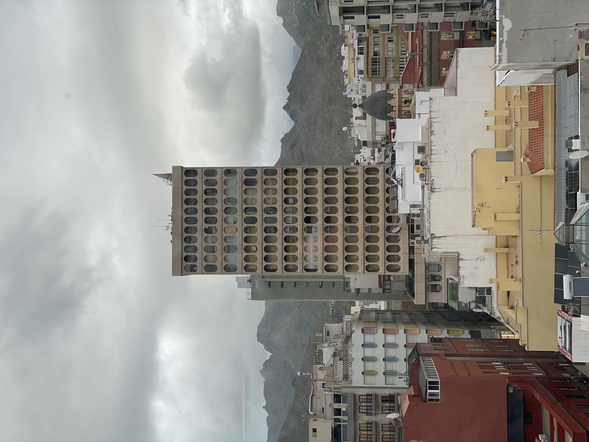 The image presents a dense urban cityscape against a backdrop of mountains and an overcast sky. A prominent, multi-story building, likely a hotel or large residential block, dominates the mid-ground on the left, characterized by a striking, repetitive facade of oval-shaped windows or openings. The building is light-colored, possibly concrete or a similar material.
Beyond this building, and stretching across the background, are rugged, rocky mountains, their peaks partially obscured by the heavy, grey clouds, indicating an overcast day.
In the foreground and to the right, a close-packed arrangement of various buildings fills the frame. These buildings feature diverse architectural styles and colors, including yellow, white, red, and beige structures. Many rooftops are visible, some equipped with solar panels, antennas, satellite dishes, and water tanks. One rooftop clearly shows the brand name "JUNKERS" on a white cylindrical tank, likely a water heater. The overall impression is one of a busy, established urban environment. There is no visible human activity, giving the scene a quiet, almost still quality despite its density.