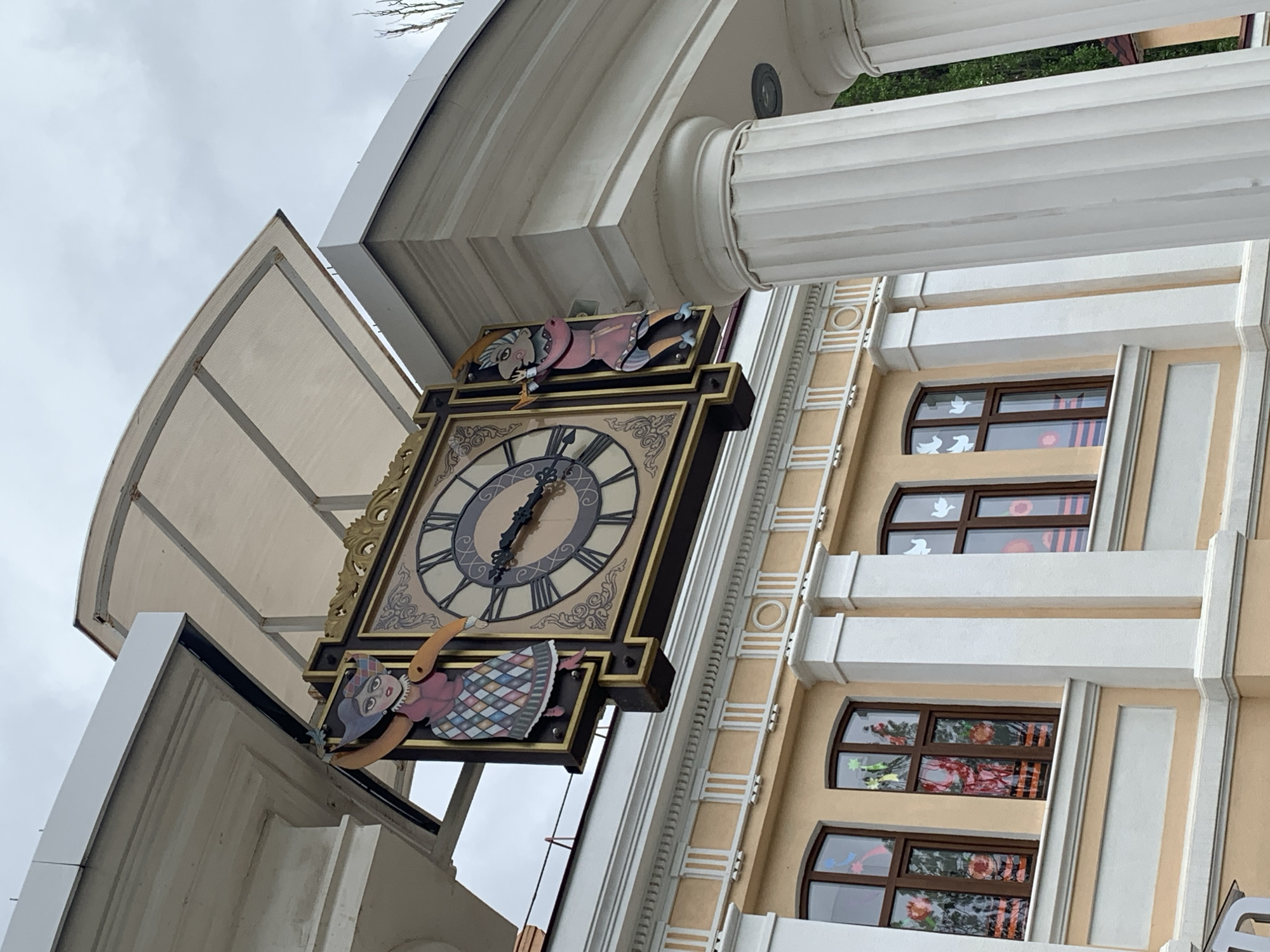 The image shows a large, decorative clock attached to a building in Stavropol, Russia. The clock face is circular with Roman numerals, and it is framed by ornate gold detailing. Cartoon figures are attached to the top and bottom of the clock. The building is multi-storied with a tan facade and white accents, including columns. The windows are arched and contain decorations. The sky is overcast.