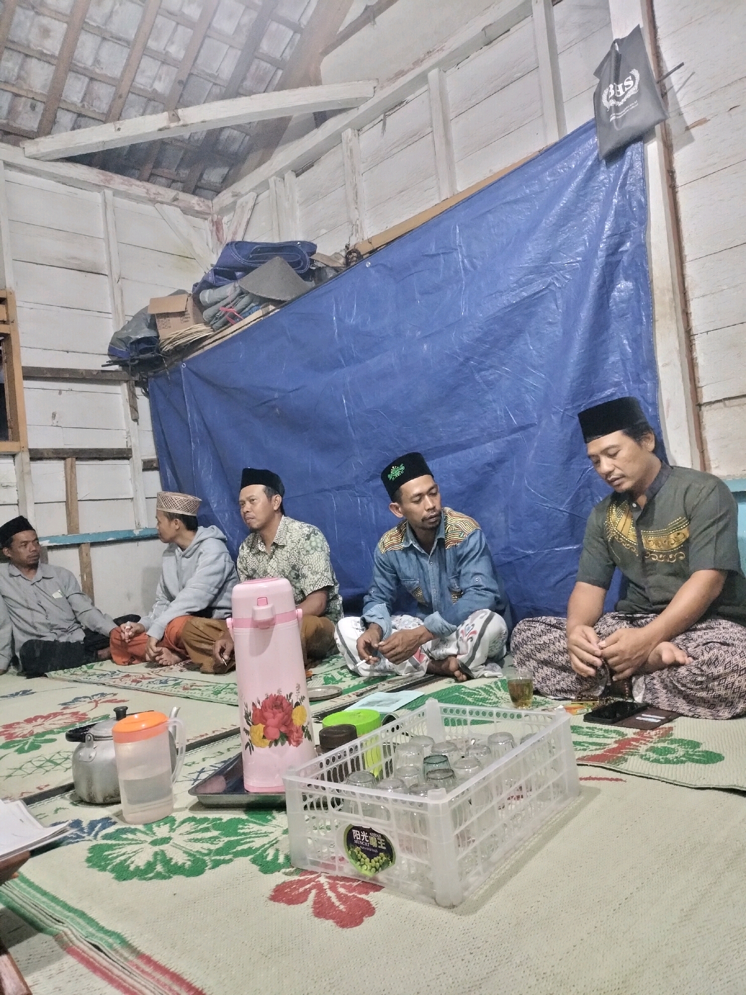 In a dimly lit, indoor space with a corrugated metal roof and unfinished wooden walls, five men are seated on a patterned mat on the floor. They are all wearing traditional Islamic clothing, including peci hats. The men appear to be engaged in a conversation or a religious gathering. In the foreground, there are various objects on the mat, including a pink thermos, a metal teapot, a pitcher of water, a tray with glasses, and a small book. A blue tarp is draped on the wall behind the men. The overall impression is one of a humble, communal gathering. The location is identified as Pujut, Indonesia.