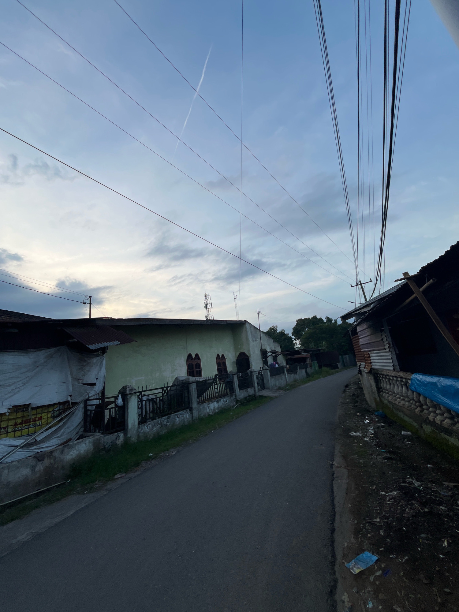 The image depicts a street scene in Patumbak Dua, Indonesia, under a twilight sky with scattered clouds and a contrail. The road, paved with asphalt, runs from the foreground into the distance, bordered on both sides by structures. On the left, a light green building with arched windows and a fence is visible, partially obscured by a white tarp. To the right, a structure with a corrugated metal roof and wooden supports is present, with a stone balustrade and blue tarp lining its base. Several utility poles with numerous overhead wires dominate the upper portion of the frame. The environment appears to be a residential or semi-urban area, with some visible debris along the roadside. No individuals are present in the scene.