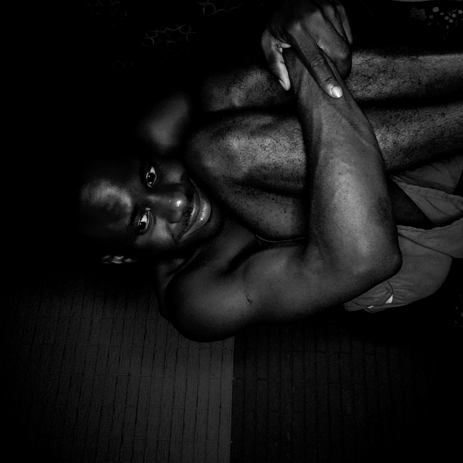 A black and white, high-angle photograph shows a shirtless black man with dark hair. The man is oriented with his head at the left side of the frame, facing upward towards the camera. His arms are folded across his chest, with one hand resting on the opposite arm. The subject appears to be sitting or lying down on a surface that is darker and appears to be made of a hard surface such as tile or linoleum. The lighting is dim, creating strong shadows and high contrast in the image. The location is not specified.