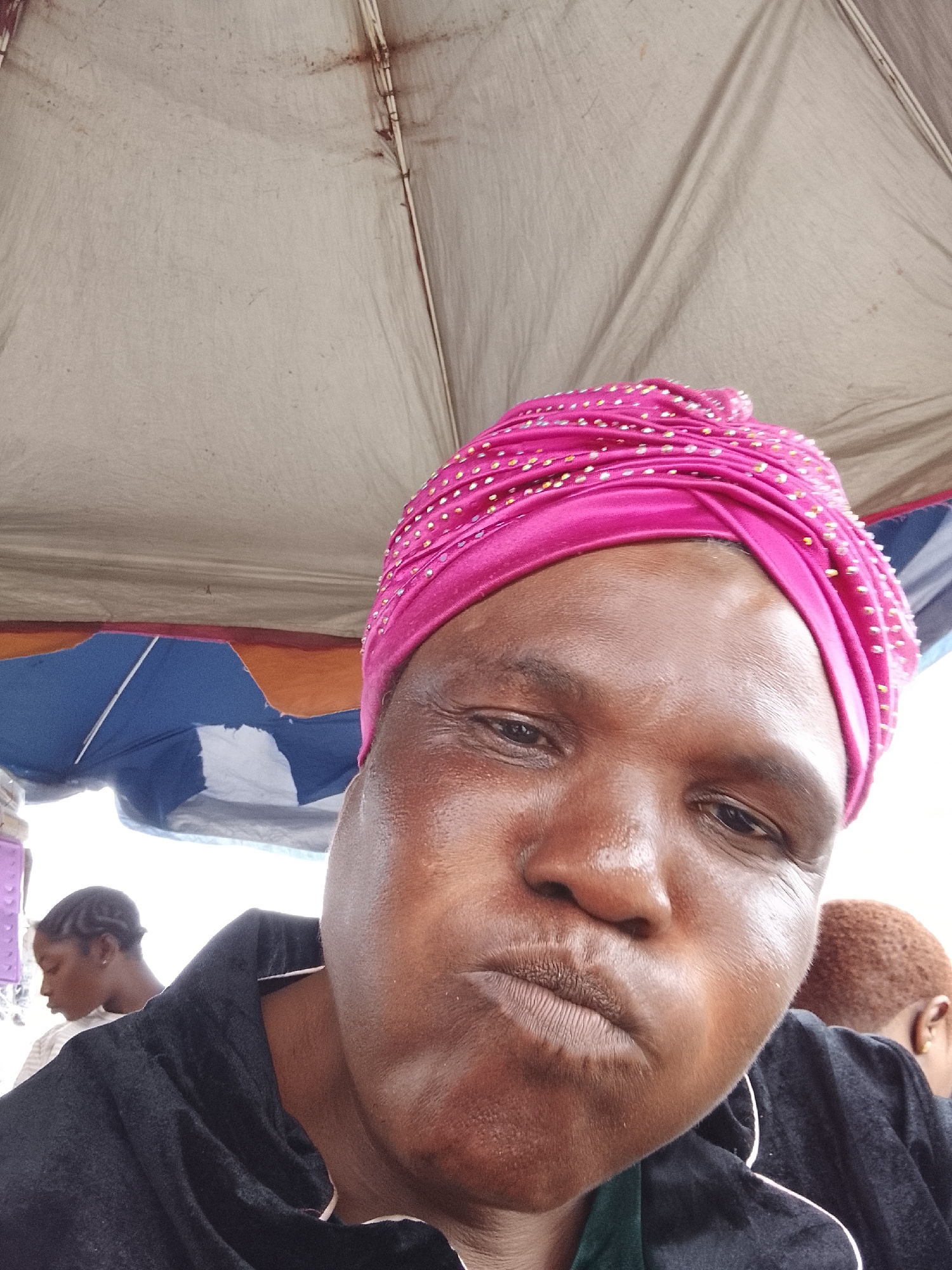 The image is a close-up selfie of a Black woman wearing a bright pink headscarf adorned with small, shiny embellishments. She has dark skin and is looking directly at the camera with her lips pursed. She is wearing a black top. In the background, to the left, another Black woman with braided hair is visible, also looking away from the camera. Further in the background, a person with reddish hair is partially visible. The scene appears to be outdoors, under a large, cream-colored tent or umbrella structure, with segments of blue and orange fabric visible behind the main subject. The overall lighting suggests it might be daytime, possibly overcast. The setting is identified as being in Lekki, Nigeria.