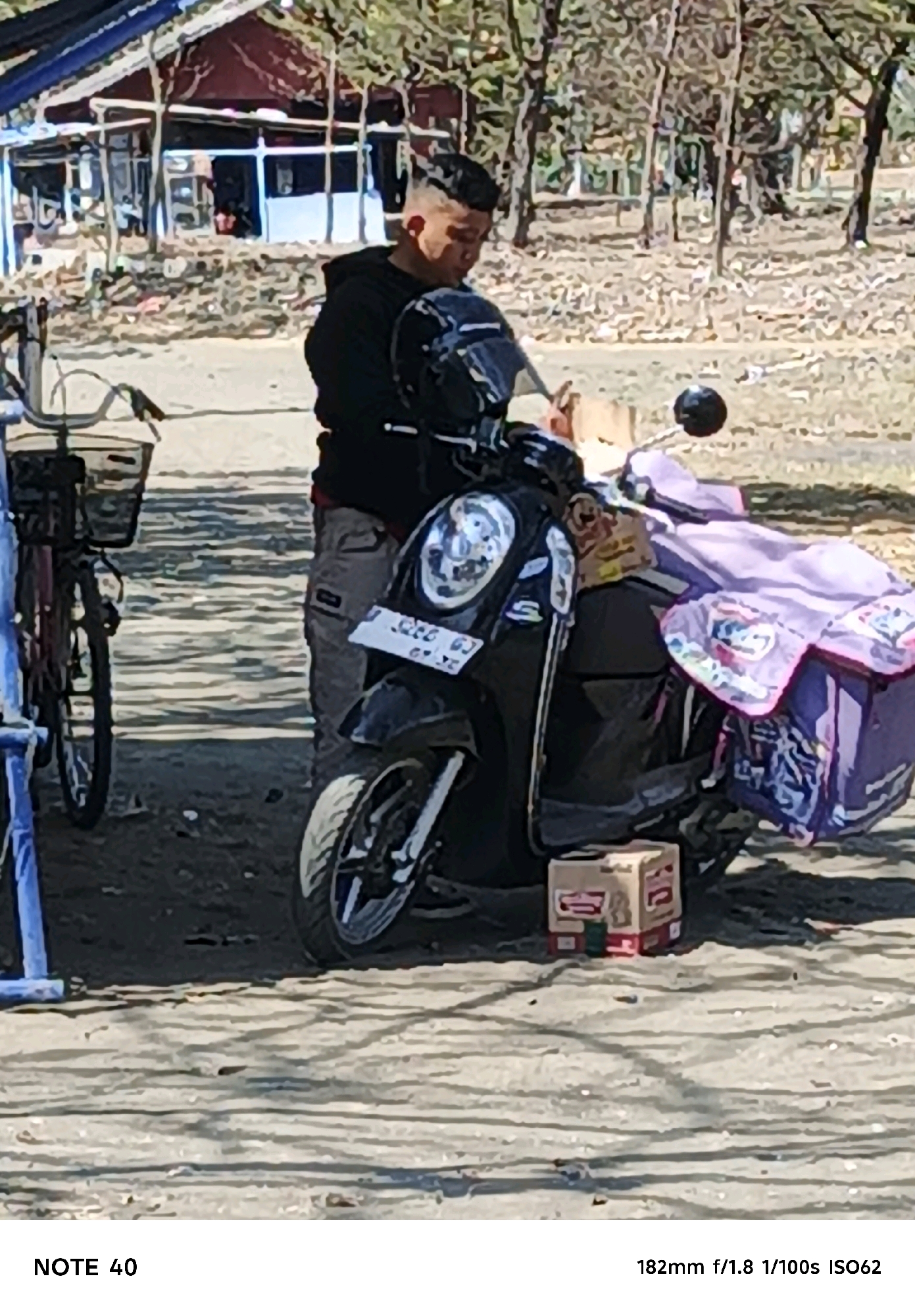 In Kelurahan Cilacap, Kecamatan Cilacap, Indonesia, a young man with a fade haircut is seated on a black scooter. He is wearing a black hoodie and grey pants. The scooter has a white license plate with red lettering. A purple bag with a patterned design is attached to the back of the scooter. In front of the scooter, a cardboard box is placed on the ground. To the left of the scooter, a bicycle with a basket is visible. The background shows a building with a blue roof and trees, suggesting an outdoor environment.
