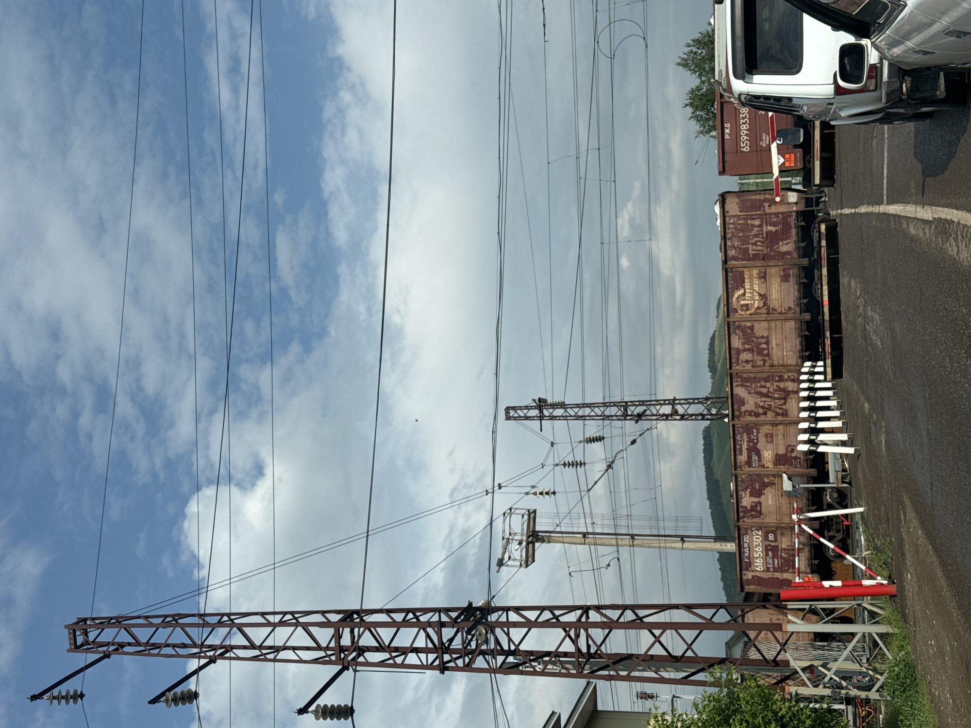 A railway crossing is depicted in Zykova, Russia, with multiple train cars stopped on the tracks. Two large, rust-colored metal towers supporting overhead electrical lines are prominent in the foreground and midground. The sky is a mix of blue with scattered white clouds. A red and white striped barrier gate is lowered across the road, indicating the train's passage. A white SUV is stopped at the crossing. The road appears to be paved with asphalt. The surrounding environment shows greenery and distant hills.