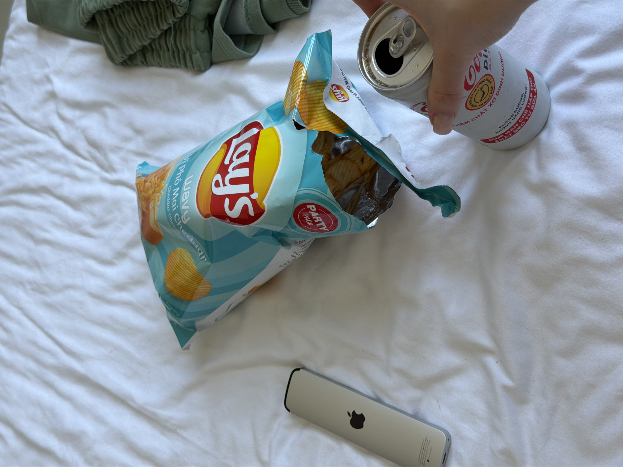 The image shows an open bag of Lay's Wavy Cheddar and Phô Mai Cheese flavored potato chips on a white bedsheet. To the right of the chip bag, a hand holds an open can of a beverage with Vietnamese text, including "KHÔNG DUONG" (No Sugar) and "KHÔNG NĂNG LƯỢNG" (No Energy). A gray Apple TV remote is visible below the chip bag. Part of a green clothing item is in the top left corner. The chips and beverage are likely being consumed. The beverage can identifies the city of Đà Nẵng, Vietnam.