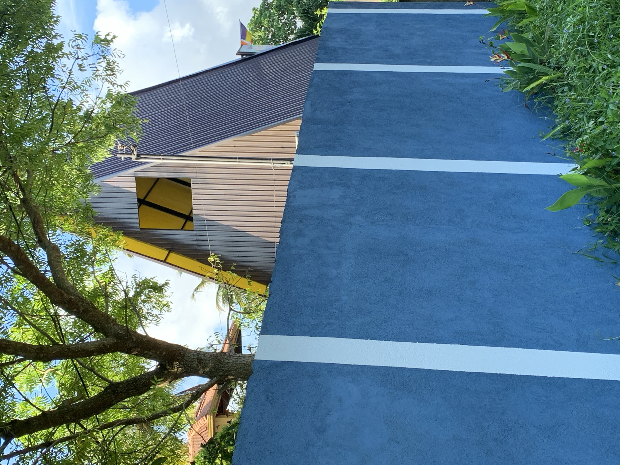 The image depicts a section of a wall with a rough, textured blue finish, adorned with three vertical white stripes. The wall is situated in Kabalana, Sri Lanka. Behind the wall, a triangular structure with a dark corrugated metal roof is visible. This structure features vertical brown wooden siding and a prominent yellow triangular window with internal bracing. A large tree with green foliage partially obscures the view of the building and the sky. The environment appears lush, with green grass and some flowering plants growing at the base of the wall. The sky is partly cloudy with a bright blue hue.