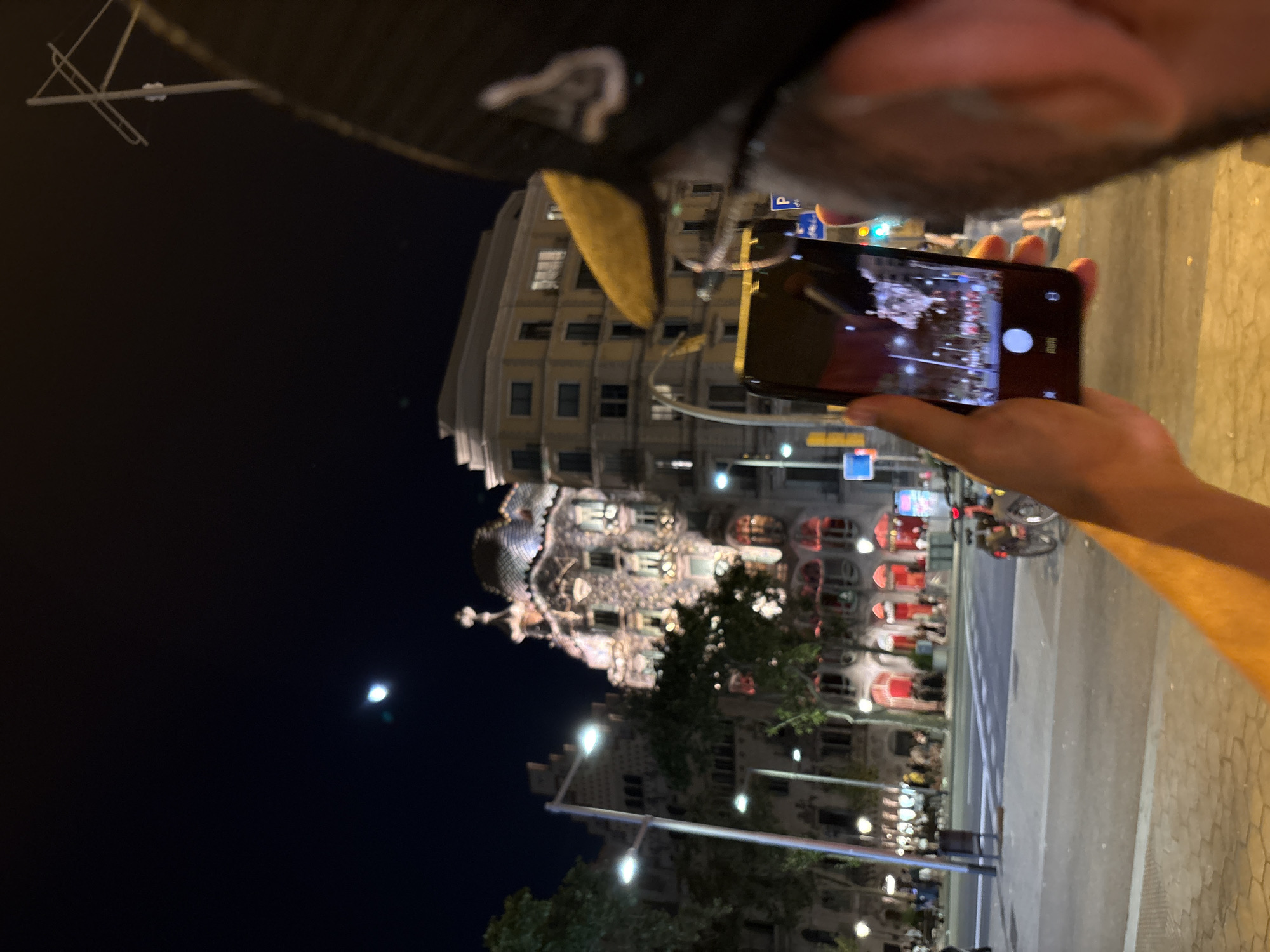 In Barcelona, Spain, at night, a person is holding a smartphone to capture a photograph of a building. The building in the background is illuminated and appears to be an ornate architectural structure with a distinct, decorated roof. The smartphone's screen displays a captured image, suggesting the user is actively photographing the scene. The foreground shows part of the person's arm and head, indicating a first-person perspective. The urban environment includes streetlights, trees, and other buildings, suggesting a public street or plaza setting. The sky is dark, with a visible moon.