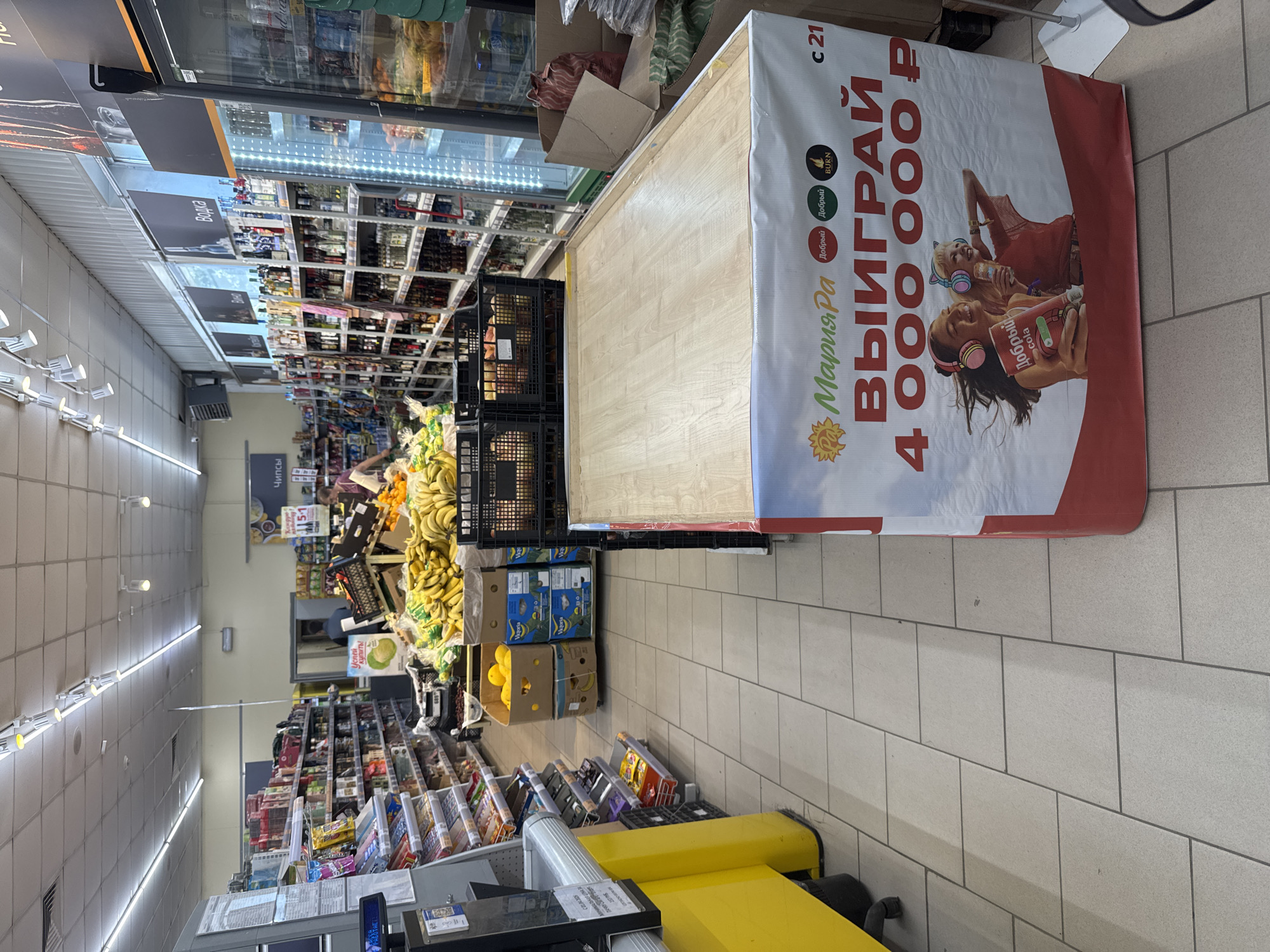 The image captures an indoor view of a supermarket in Soloneshnoye, Russia. The aisle is lined with shelves stocked with various products, including snacks, drinks, and other groceries. In the foreground, a promotional display for "Maria-Ra" is visible, featuring an advertisement for winning 4,000,000 rubles. Two young women wearing headphones are depicted on the advertisement. To the left, a checkout counter with a yellow front is partially visible. The floor is tiled with light-colored squares. The overall scene depicts a typical retail environment with an emphasis on a promotional campaign.