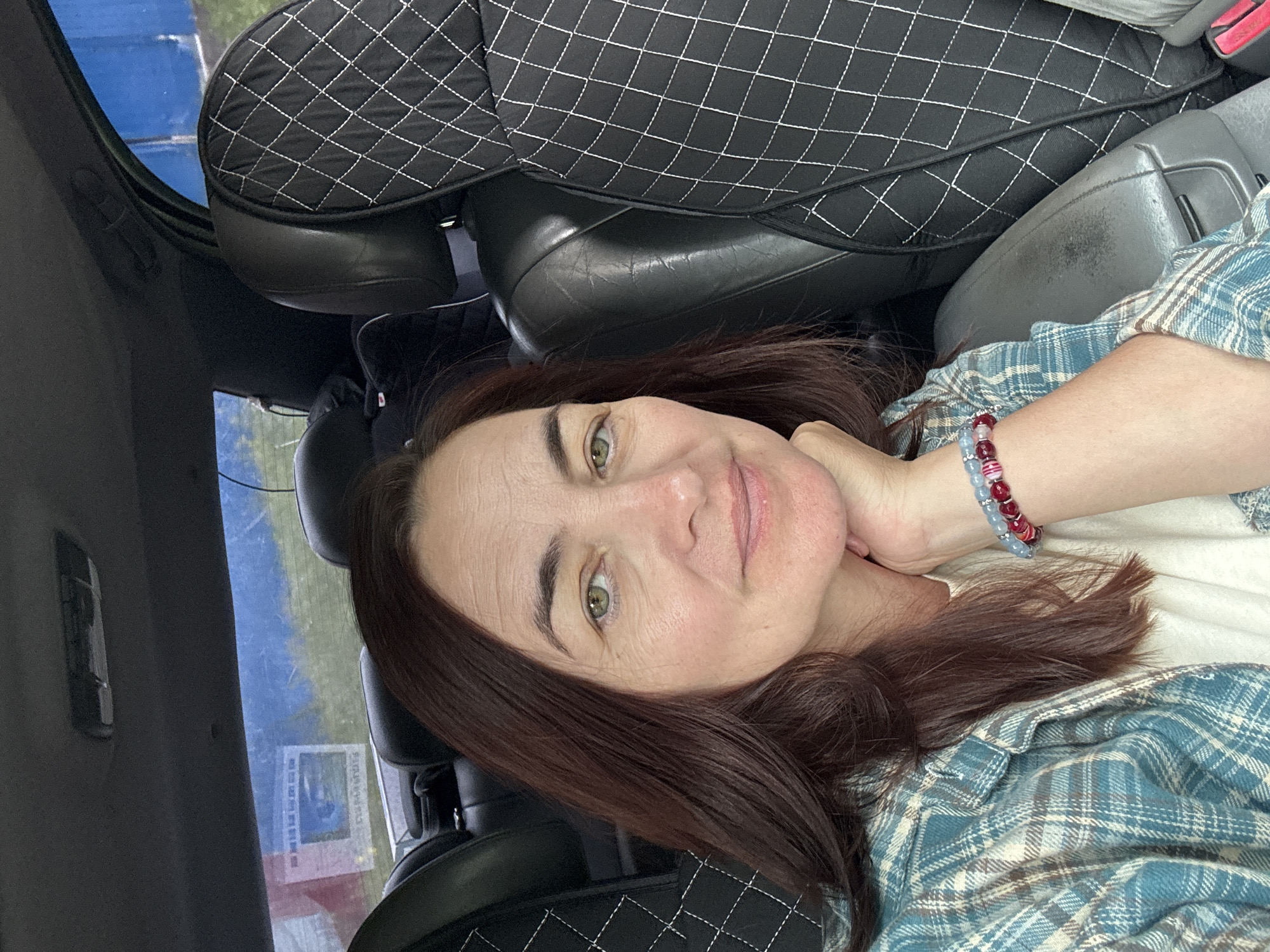 The image shows a close-up, selfie-style portrait of a woman with brown hair, seated in the passenger seat of a car. She is wearing a blue and white plaid shirt over a cream-colored top, and a bracelet on her left wrist. The woman is resting her chin on her hand and looking towards the camera with a slight smile. The background includes the interior of the car, with black leather seats and a headrest cover with a diamond pattern. Through the rear window, a blue tarp or banner and some greenery are visible. Based on context, the car is likely in Ufa, Russia, given the provided location.