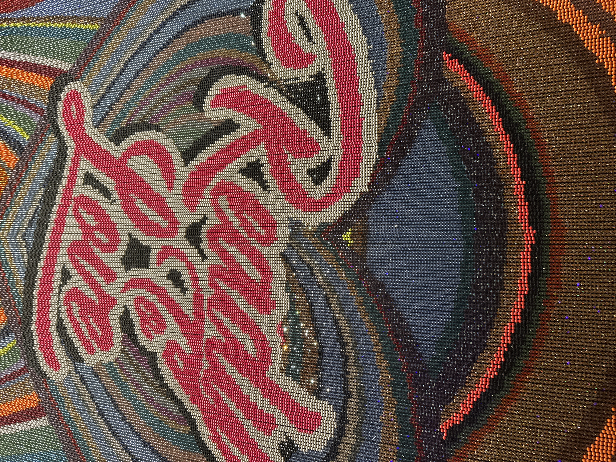 The image is a close-up, overhead shot of a colorful, beaded tapestry or artwork featuring the words "Ready for Love" in large, stylized pink lettering with white and black outlines. The lettering is centered within concentric circles of various colors, including blue, gray, green, brown, and orange, all formed by small beads. The beads themselves appear to have a slight shimmer or sparkle. This artwork is part of the Swarovksi Kristallwelten (Crystal Worlds) in Wattens, Austria, specifically located in the "Great Hall" where it is a permanent installation. There are no people or actions depicted in the image.