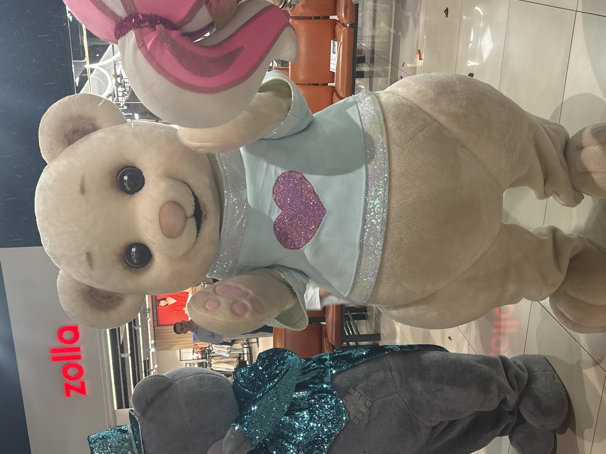 A photograph taken in the Tri Protoka, Russia area shows two plush animal mascots standing near a “Zolla” store. The first mascot is a tan-colored bear wearing a light blue top decorated with a sparkling heart and a sparkling white trim. It holds a large pink-and-white decorative item, possibly a sweet treat, aloft. A second, smaller gray bear with blue sequined clothing stands at the base of the picture. Clothing racks are visible within the store, with shoppers browsing clothing.