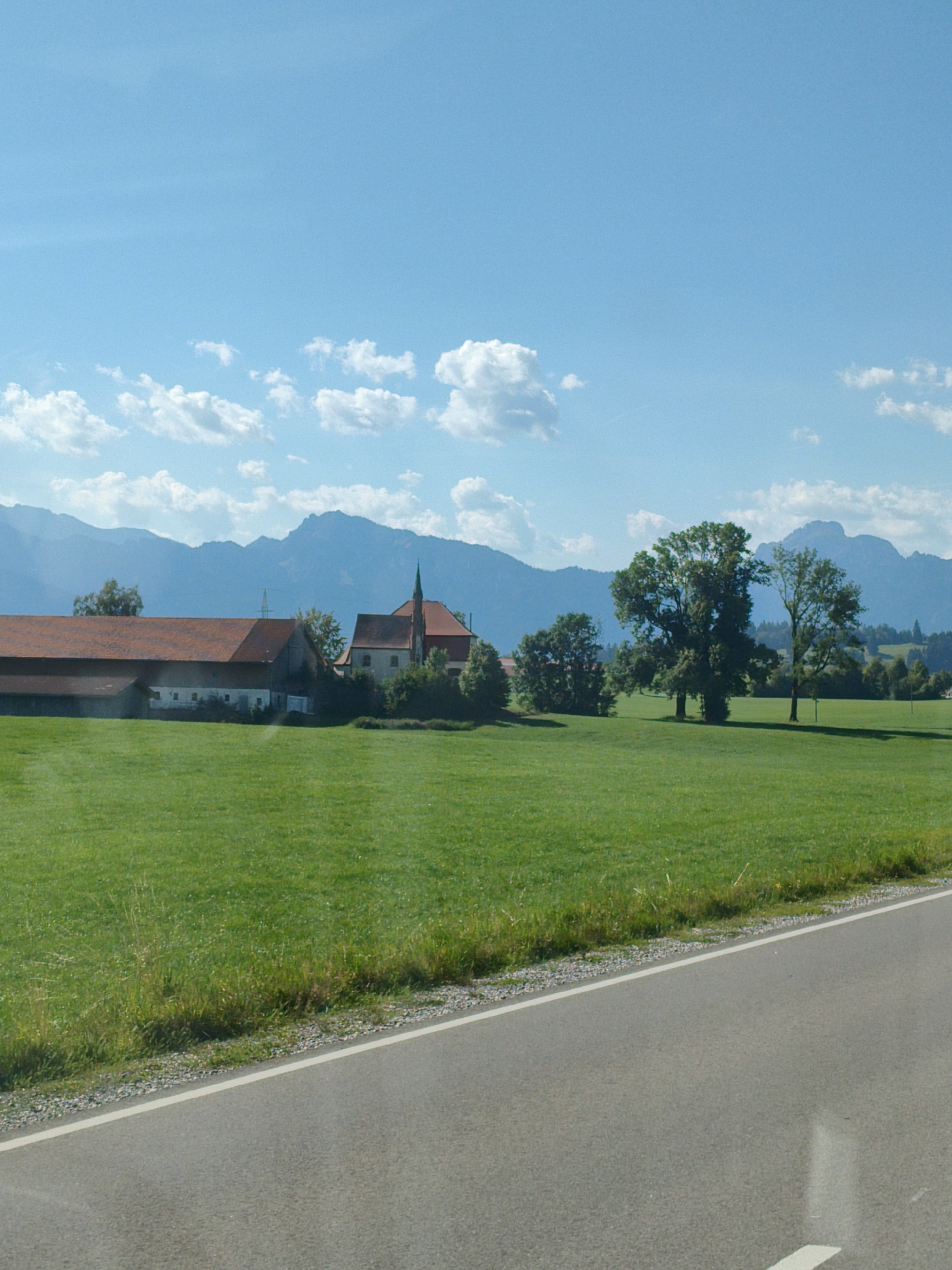 The image depicts a pastoral landscape in Roßhaupten, Germany, characterized by a clear blue sky with scattered white clouds. In the mid-ground, a sprawling farm complex with red tiled roofs is visible, featuring multiple buildings. Adjacent to the farm is a church with a prominent steeple. The foreground is dominated by a vibrant green meadow, which transitions to a paved road on the right side of the frame. In the background, a range of majestic blue-tinged mountains forms a dramatic backdrop to the scene, with lush green trees dotting the landscape between the farm and the mountains. No people or animals are present in the image, and the scene appears tranquil and undisturbed. The lighting suggests a sunny day.