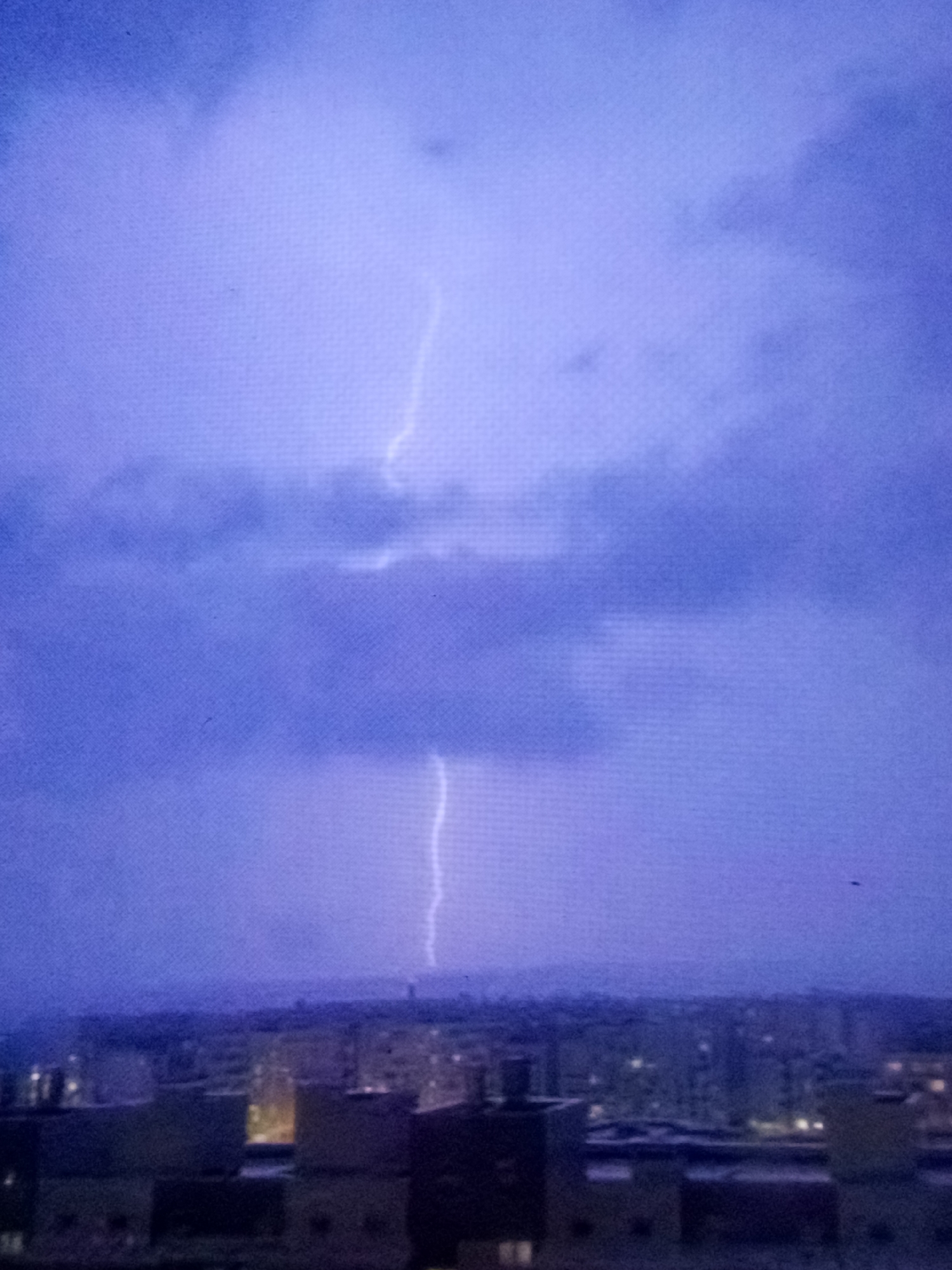A vertical lightning bolt illuminates a city skyline at dusk. The sky is a deep purple, filled with ominous, dark clouds. The city below is a blur of buildings with some windows lit, suggesting nighttime. The lightning bolt appears to strike from the upper half of the frame downwards, creating a stark contrast against the darkened sky.