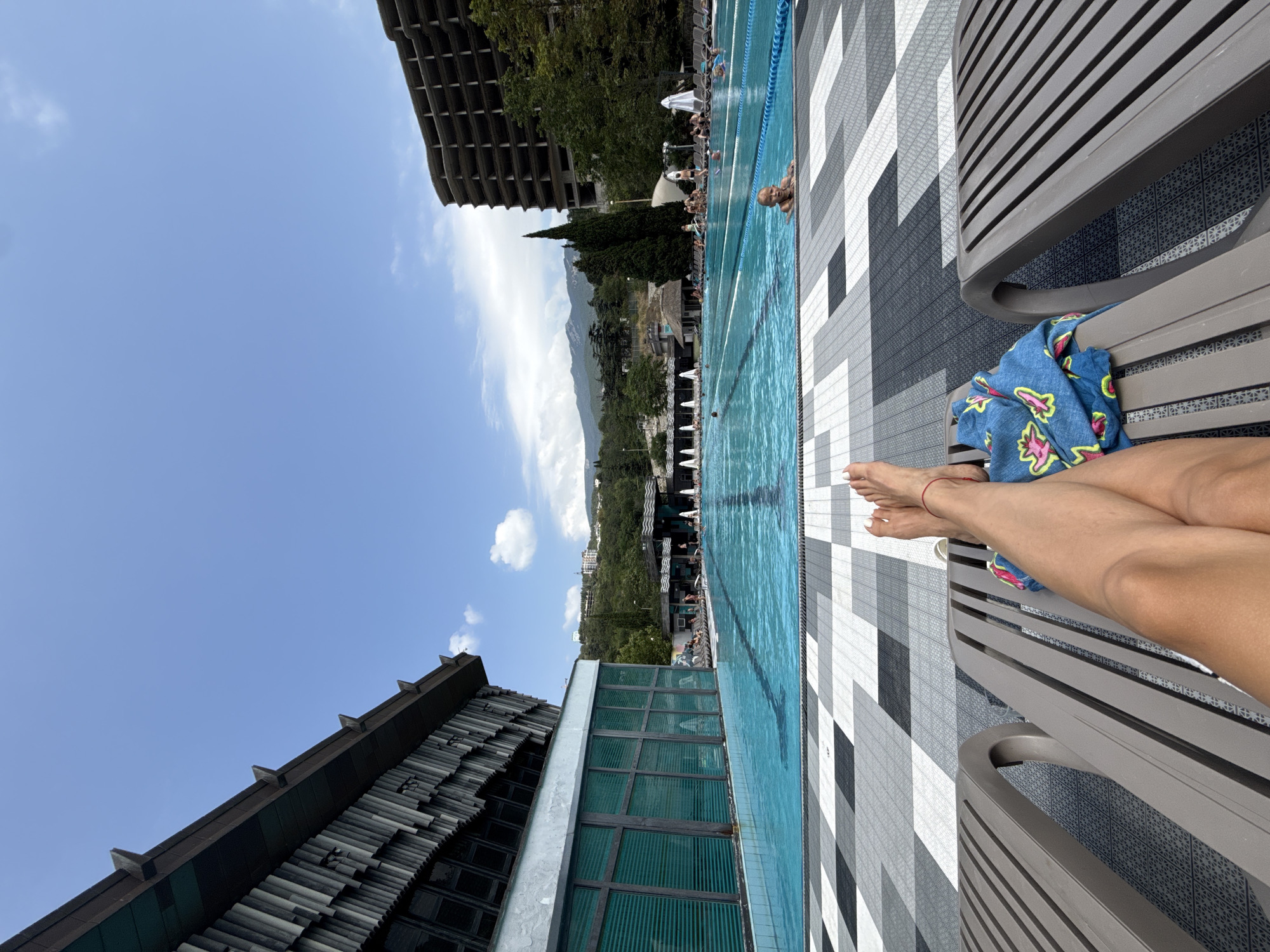 The image depicts an outdoor swimming pool area in Yalta, Ukraine. In the foreground, a person's legs are resting on a lounge chair, with a patterned towel draped over the chair. The pool is large and filled with blue water, marked with lanes. Several people are visible in and around the pool, some swimming and others relaxing on lounge chairs. To the left, a modernist building with a distinctive facade and large windows borders the pool. In the background, there are more buildings, trees, and mountainous terrain. The sky is clear and blue with some scattered clouds.